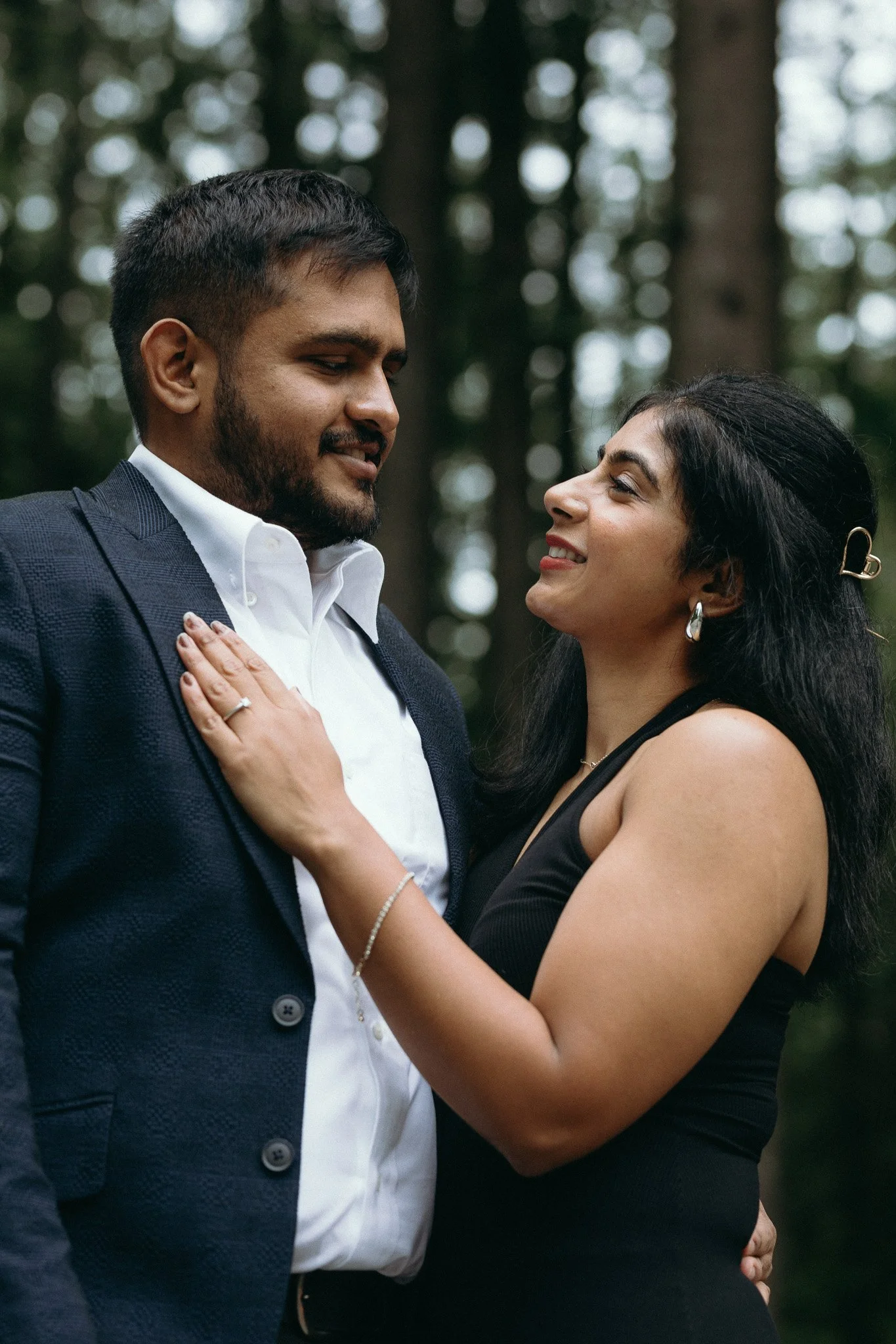 A couple sharing an intimate moment in a forest, with the woman touching the man's chest and smiling up at him.