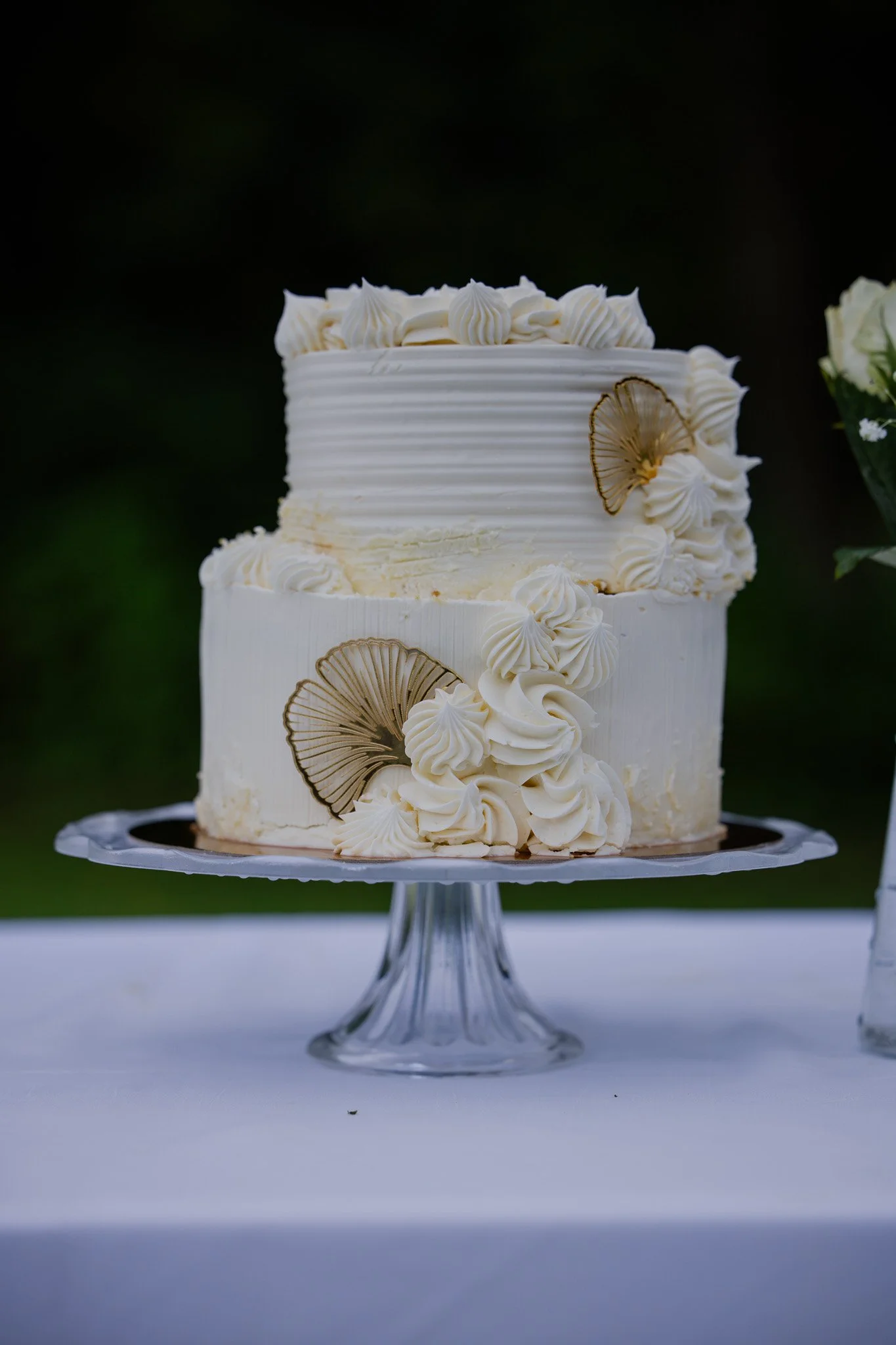 A white frosted cake decorated with vanilla cream swirls and delicate gold-colored fans, placed on a glass cake stand on a white table, with a blurred green outdoor background.
