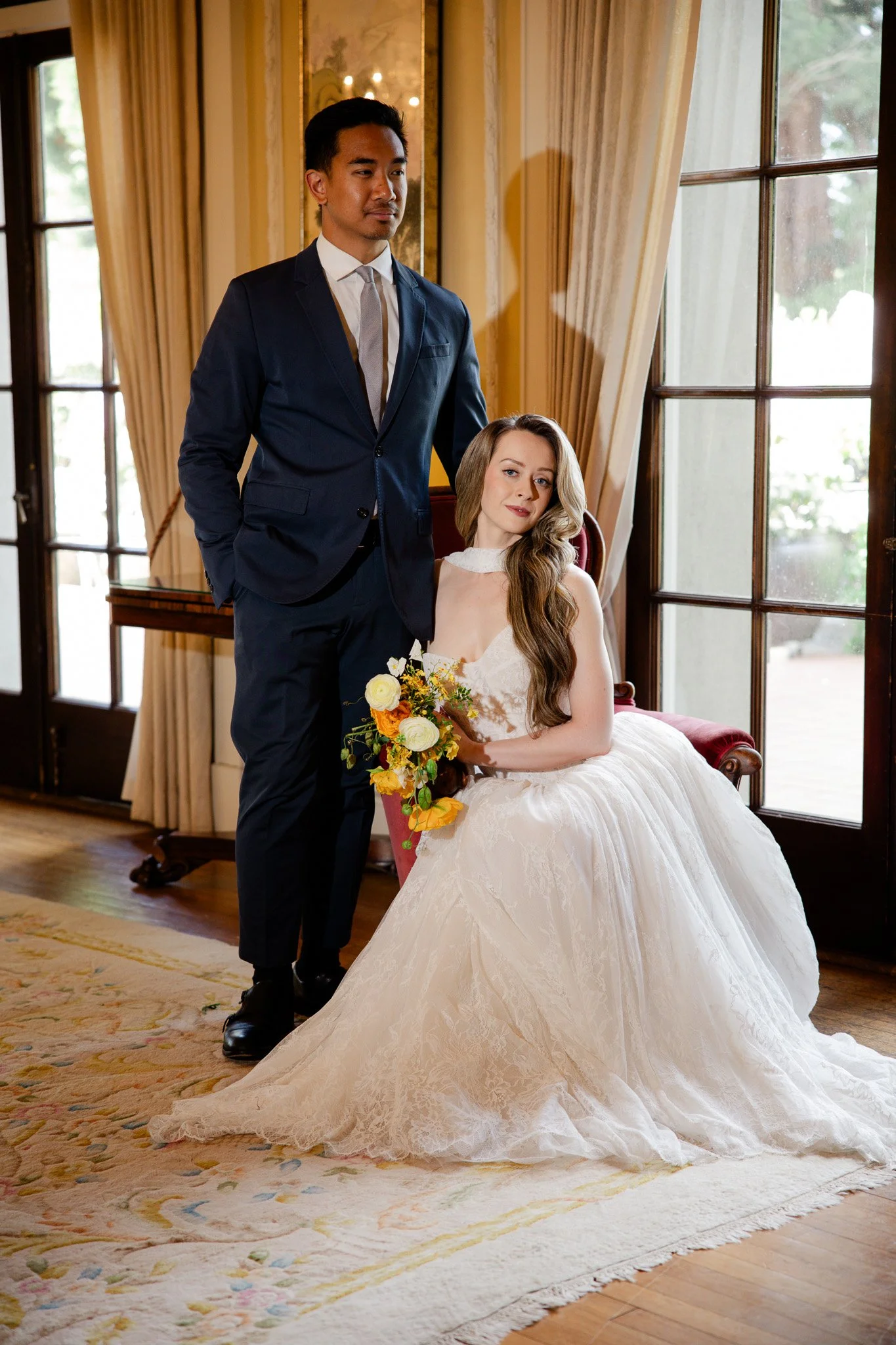 A bride in a white wedding gown holding a bouquet of yellow and white flowers sitting on a red velvet chair, with a groom in a navy suit standing beside her in a room with large windows, yellow curtains, and a patterned rug.