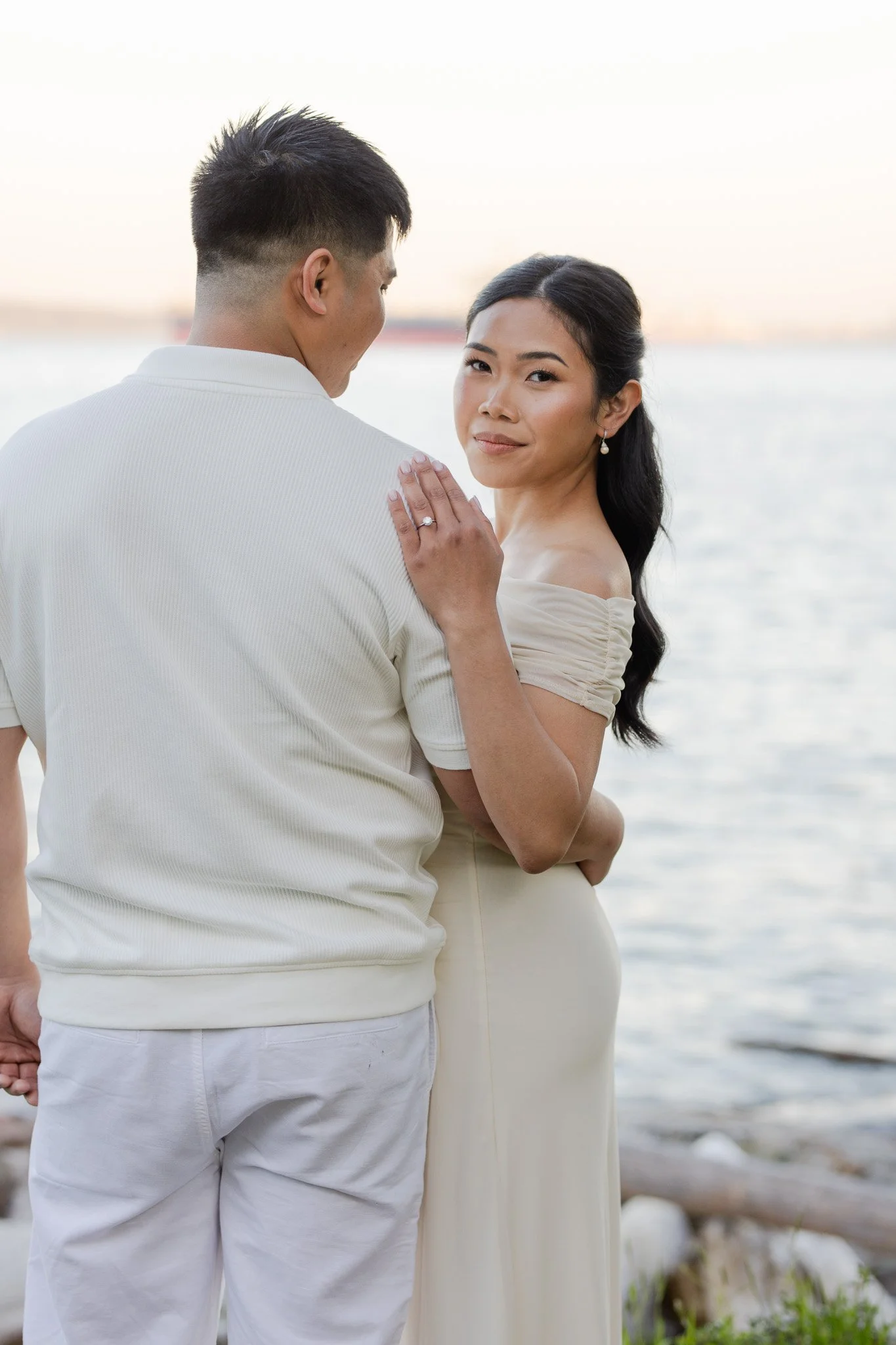 A couple standing by the water, with the woman showing her wedding ring and wearing an off-shoulder dress, while the man, dressed in white, looks at her.