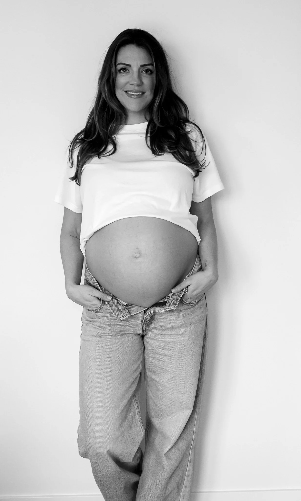A pregnant woman with long hair standing against a plain wall, wearing a cropped t-shirt and jeans, smiling at the camera.