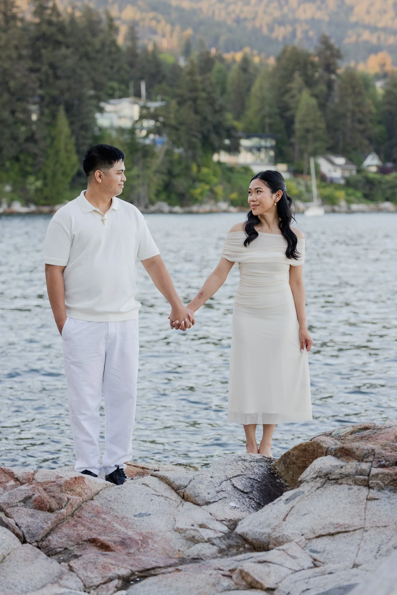 A young couple holding hands and standing on rocks by a body of water with shoreline houses and trees in the background.