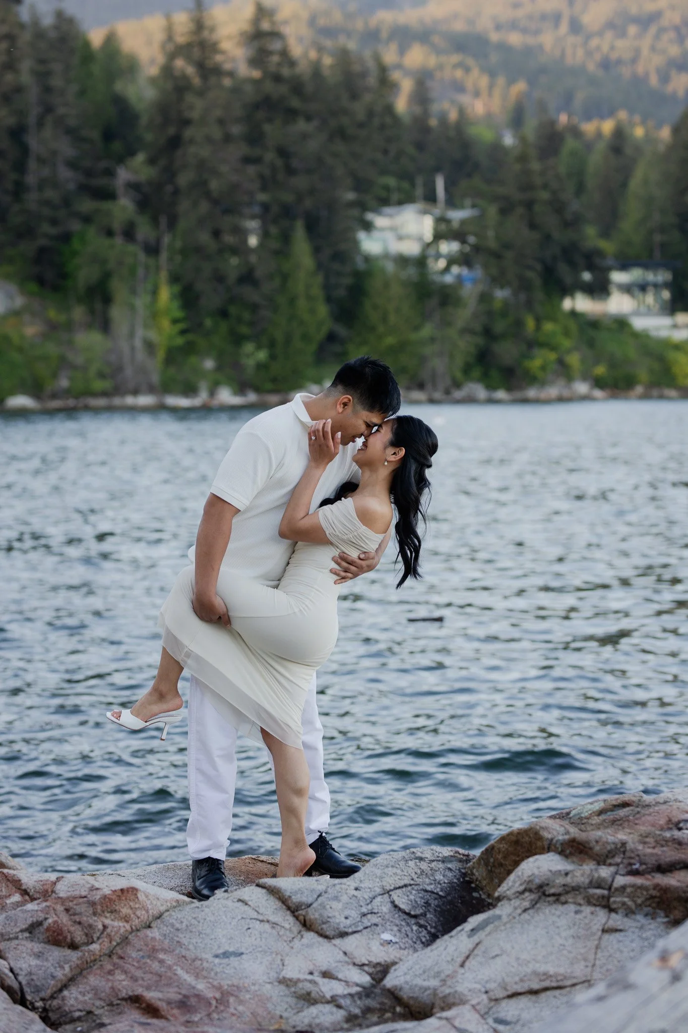 A couple by a body of water with mountains and trees in the background. The man is holding the woman in a dip, and they are smiling at each other.