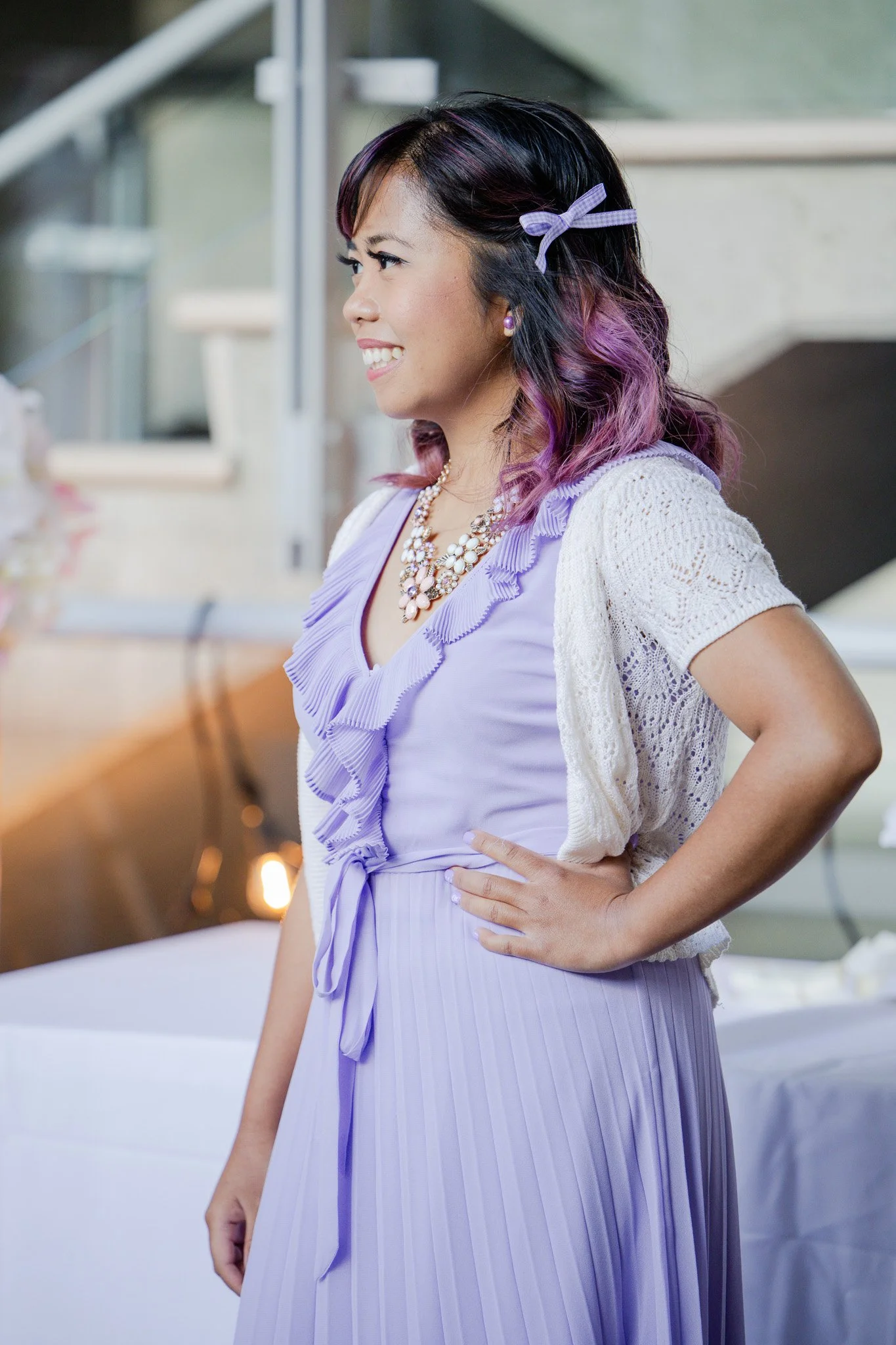 A woman with black and purple hair, wearing a lavender dress with ruffles and a bow, accessorized with a pearl necklace, earrings, and a lavender bow in her hair. She is standing indoors with her hand on her hip, smiling, and looking to her left.