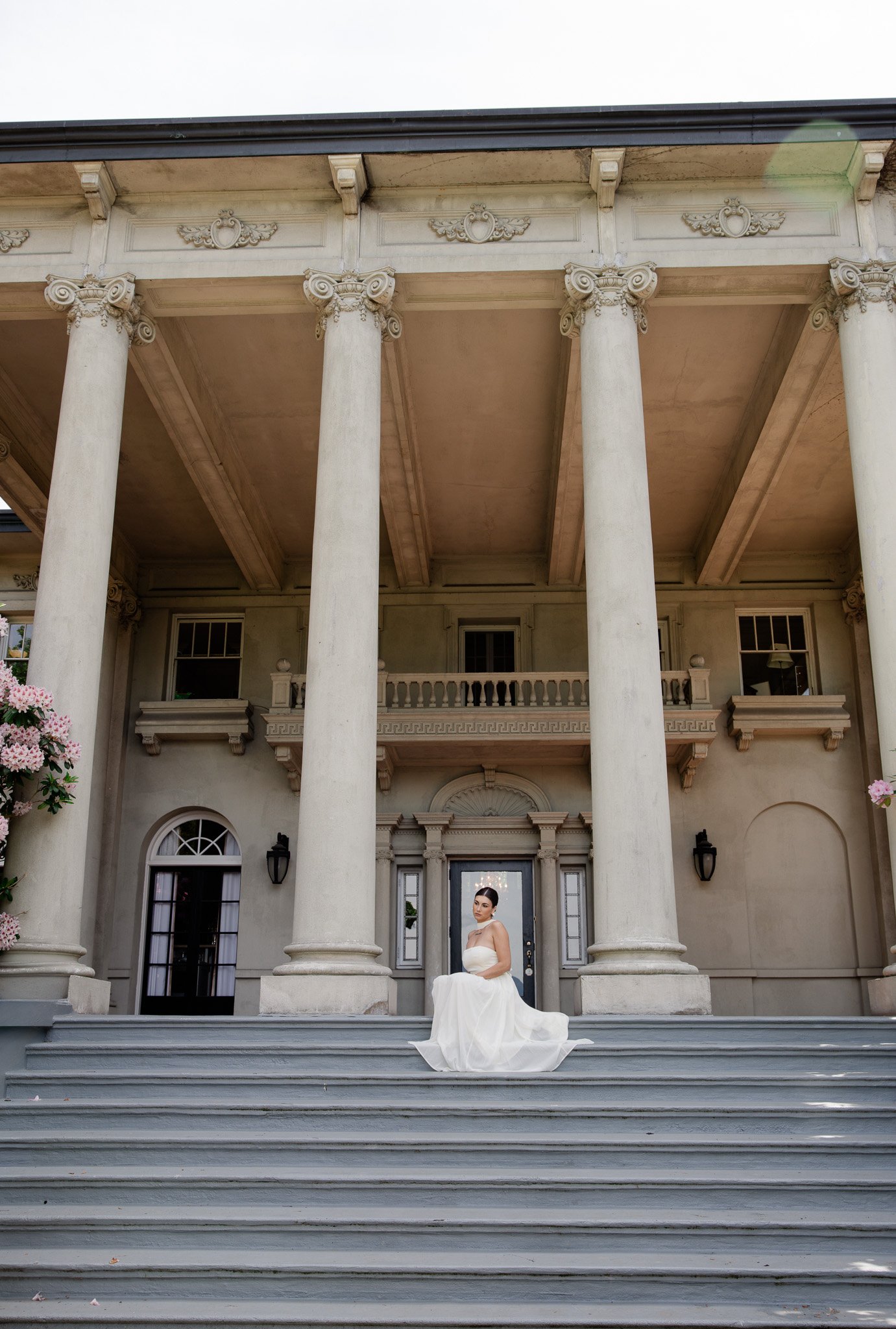 A woman in a white strapless dress sitting on the steps of a grand, classical building with large columns and ornate architectural details.