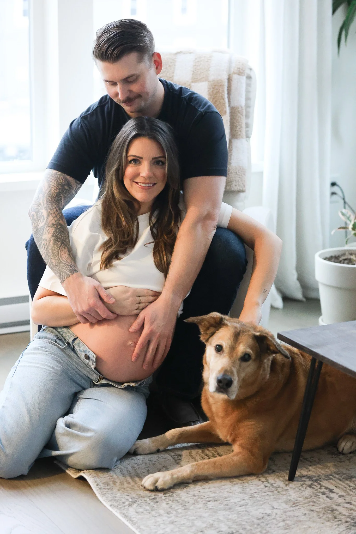A pregnant woman with long brown hair, sitting on the floor, smiling at the camera, with a man behind her gently pressing his hands on her belly. There is a dog lying beside her, resting on a rug, in a cozy indoor setting with plants and natural ligh