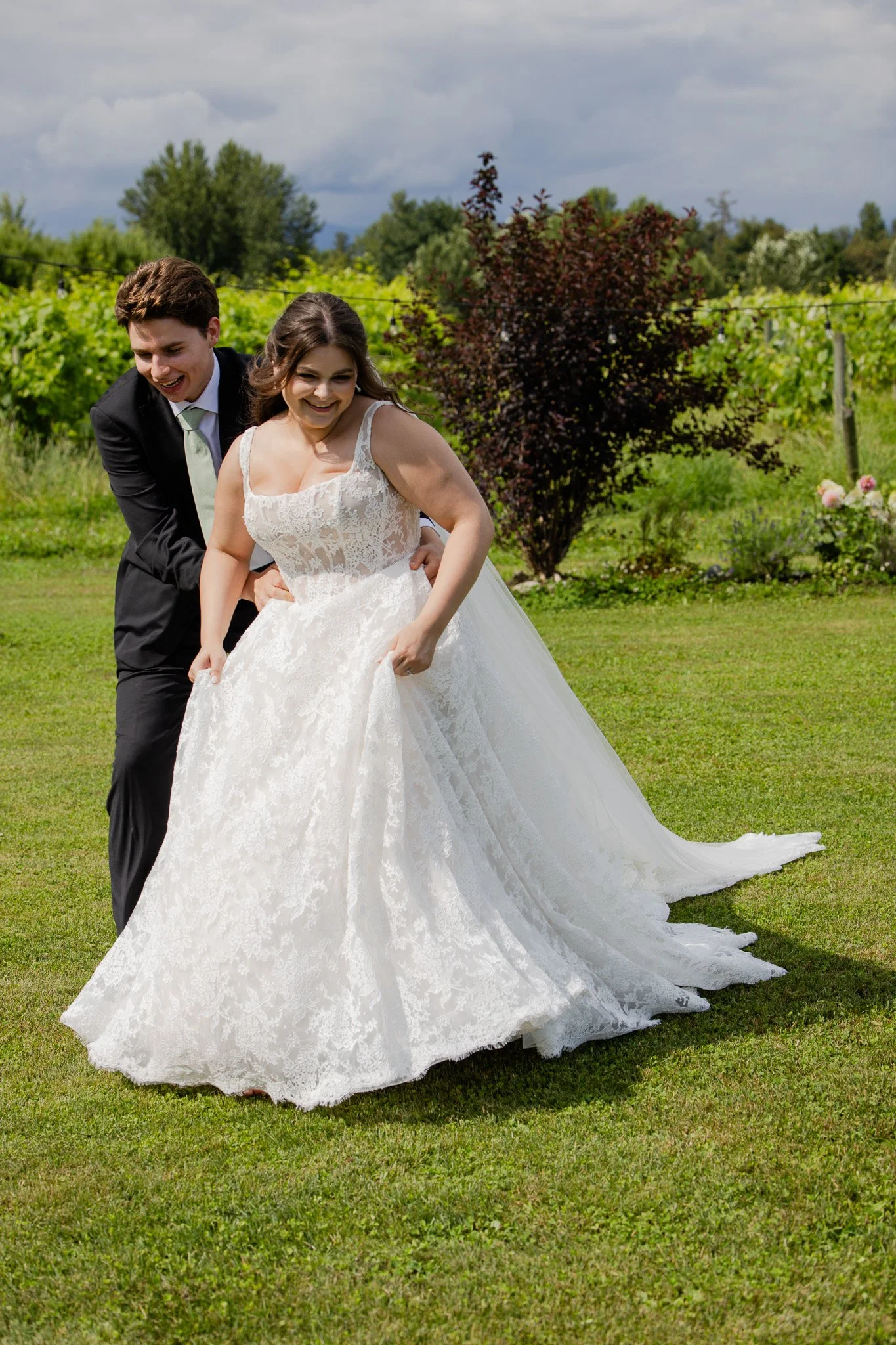 A young bride and groom celebrating outdoors on a grassy area, with trees and dark clouds in the background. The bride is in a white lace wedding gown and the groom is in a dark suit with a light tie, both appearing joyful and happy.