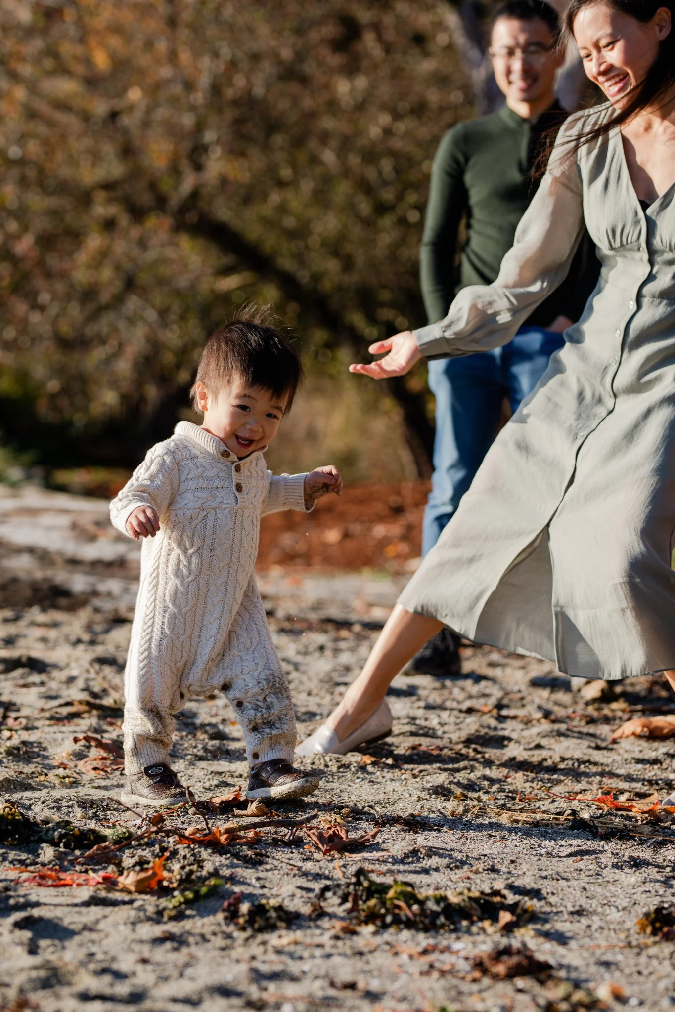 A young child in a cream-colored sweater walking on a sandy beach with fallen leaves, smiling, with two adults in the background, one woman in a gray dress reaching out to the child, and a man in a green shirt and glasses standing behind her, all out