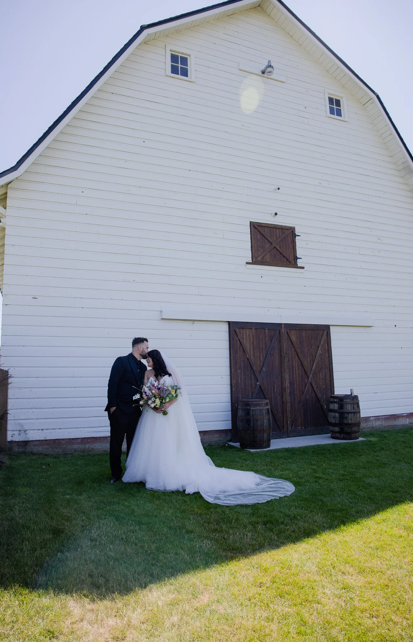Couple dressed in wedding attire standing on grass in front of a large white barn with wooden doors and barrels, sharing a kiss.
