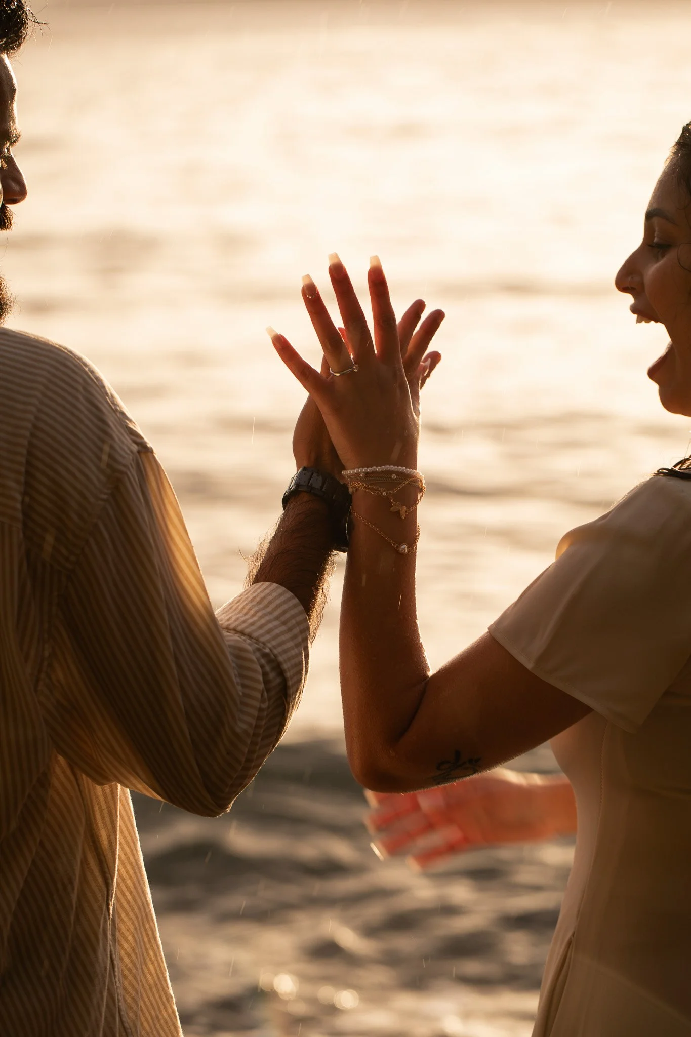 A man and woman holding hands and touching palms near the water at sunset, smiling and enjoying a romantic moment.