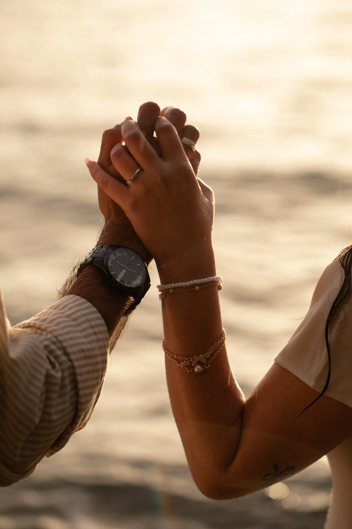 A couple holding hands with intertwined fingers near the water at sunset, wearing wedding rings and jewelry.