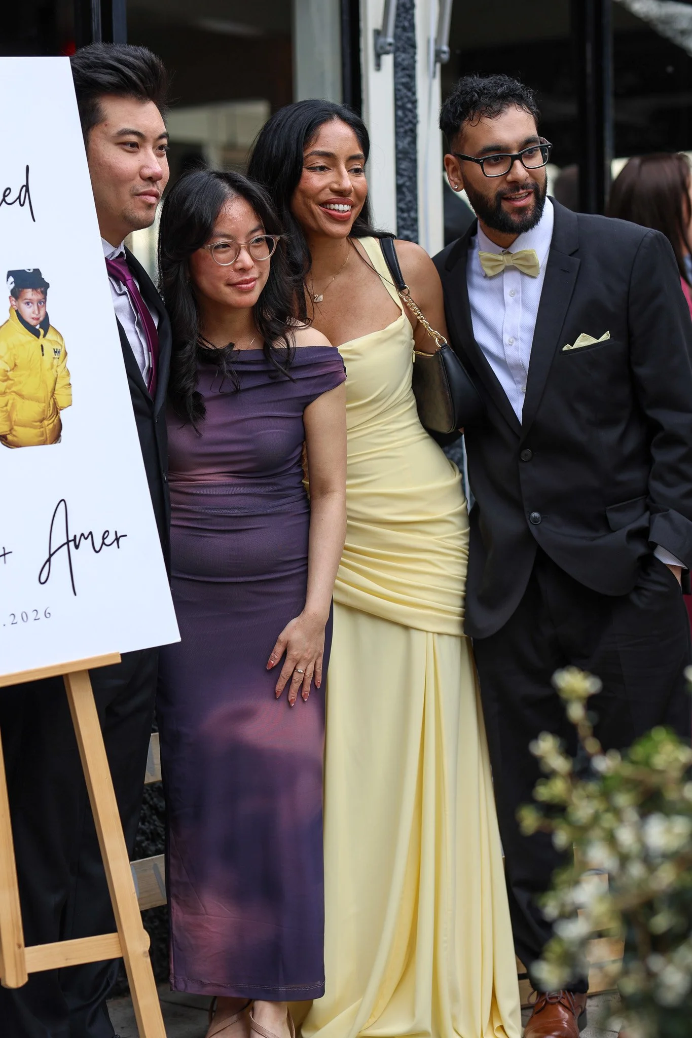 A group of four diverse people at a formal event, posing for a photo in front of a sign. They are dressed in elegant attire, smiling and appearing happy.