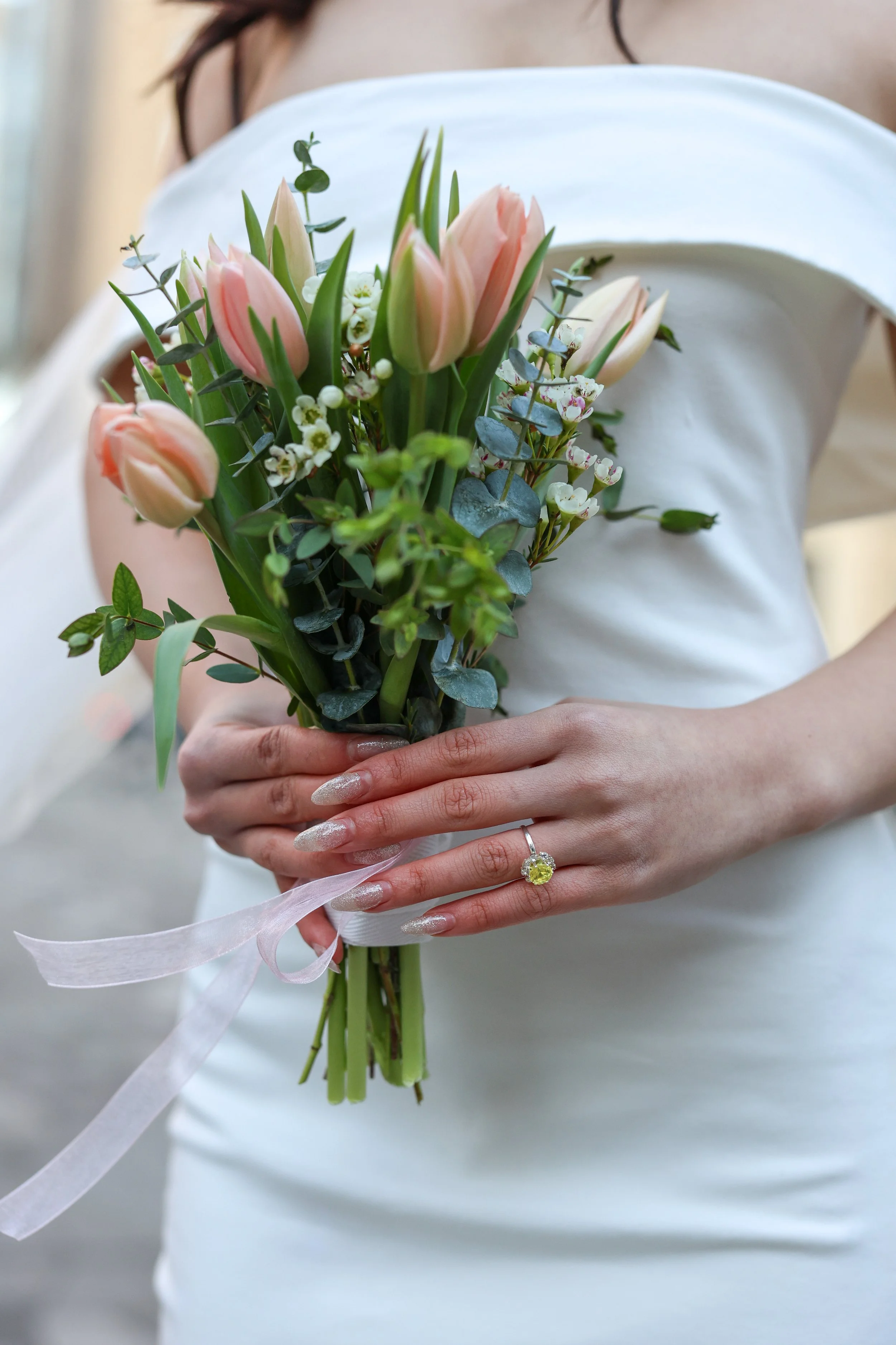A woman in a white dress holds a bouquet of pink tulips and small white flowers, with a ring on her finger.
