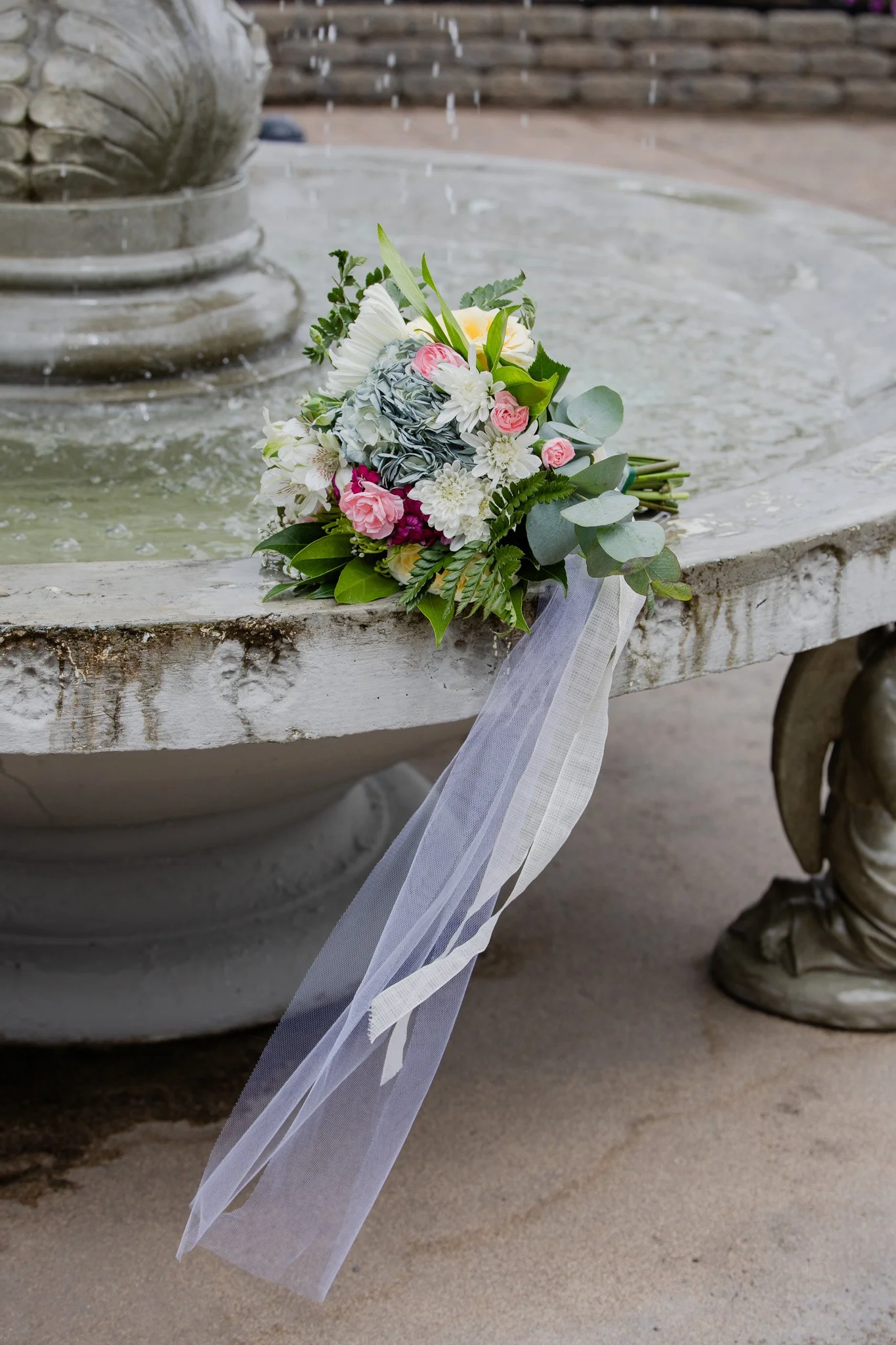 Bouquet of mixed white and pink flowers with greenery, resting on the edge of a weathered stone fountain with water and a decorative base, outdoors with sandy ground.