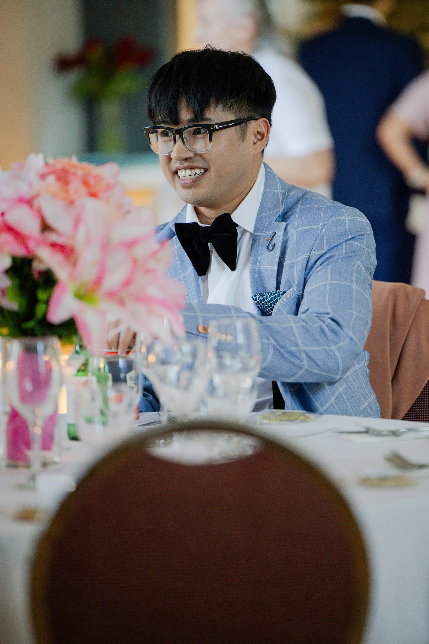 A man with black hair, glasses, and a blue checkered suit with a black bow tie smiling at a social event or gathering. There are pink flowers and glasses on a table in the foreground, with people in formal attire in the background.