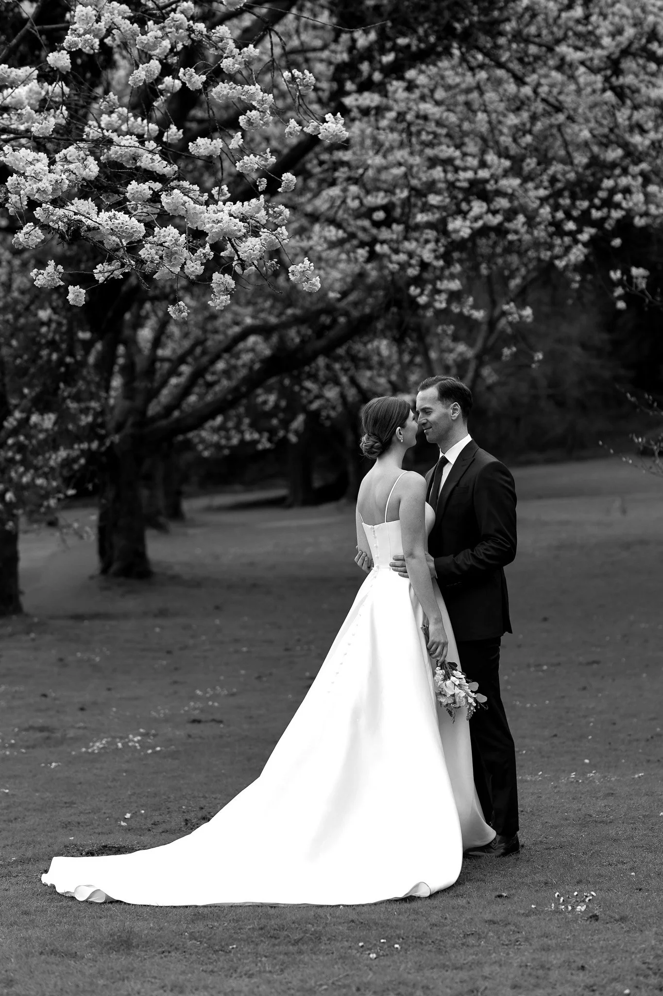 A black-and-white photograph of a bride and groom standing close together outdoors under blooming trees. The bride is wearing a strapless wedding gown with a long train, and the groom is in a dark suit. They are looking into each other's eyes and hol
