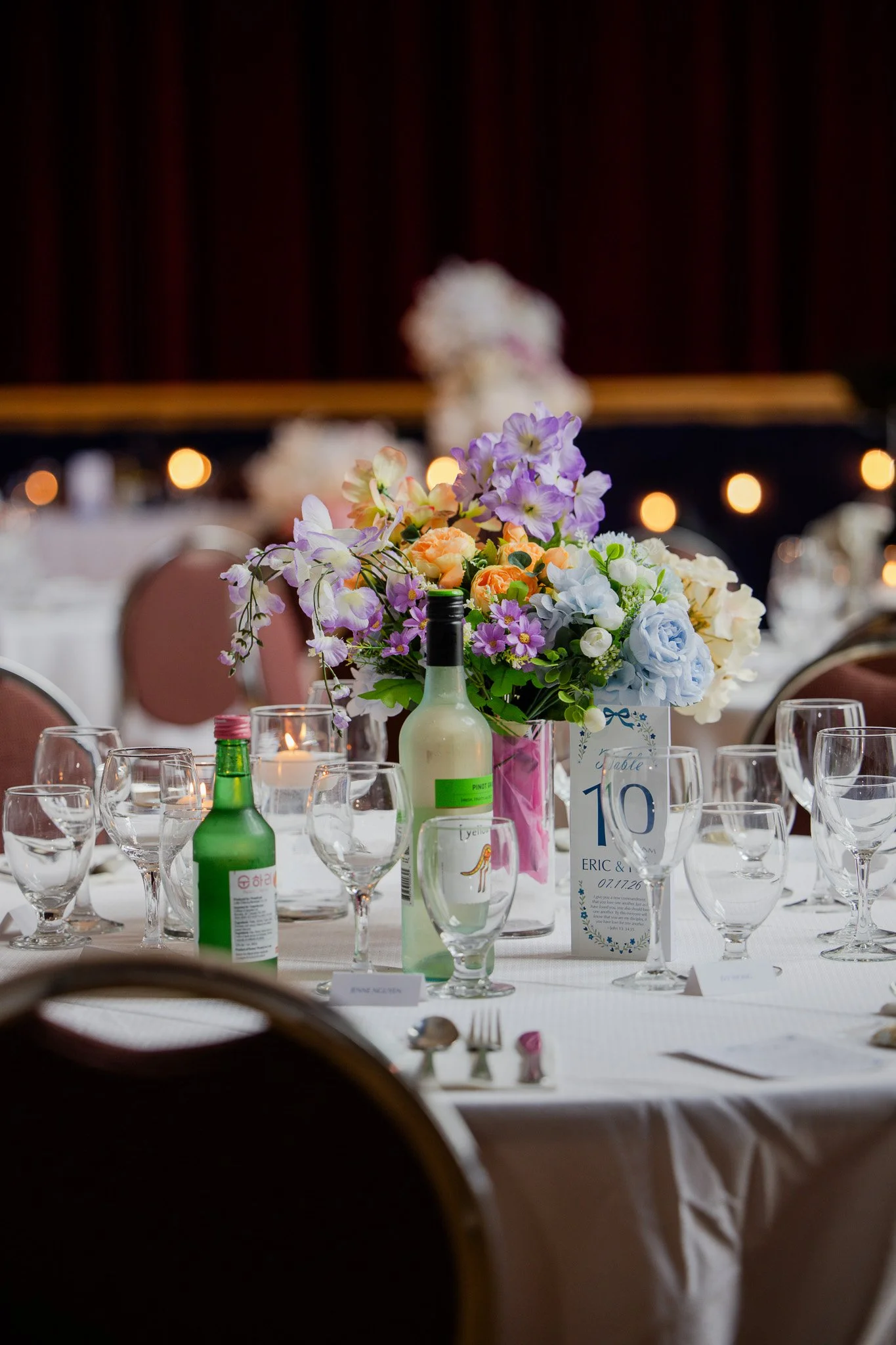 A banquet table decorated with a colorful flower arrangement, wine bottles, glasses, and place cards at a formal event or wedding reception.