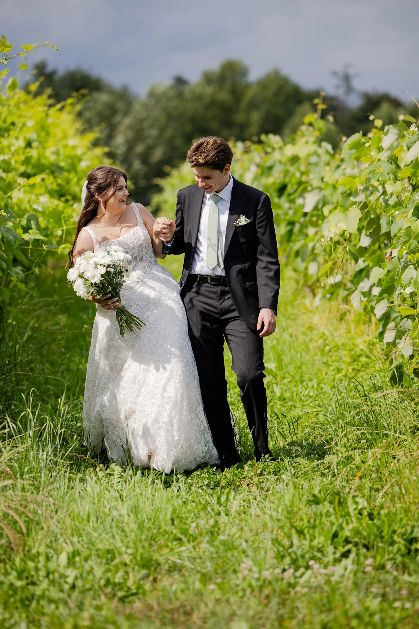 A bride and groom smiling and holding hands in a green outdoor setting on a sunny day, surrounded by lush foliage.