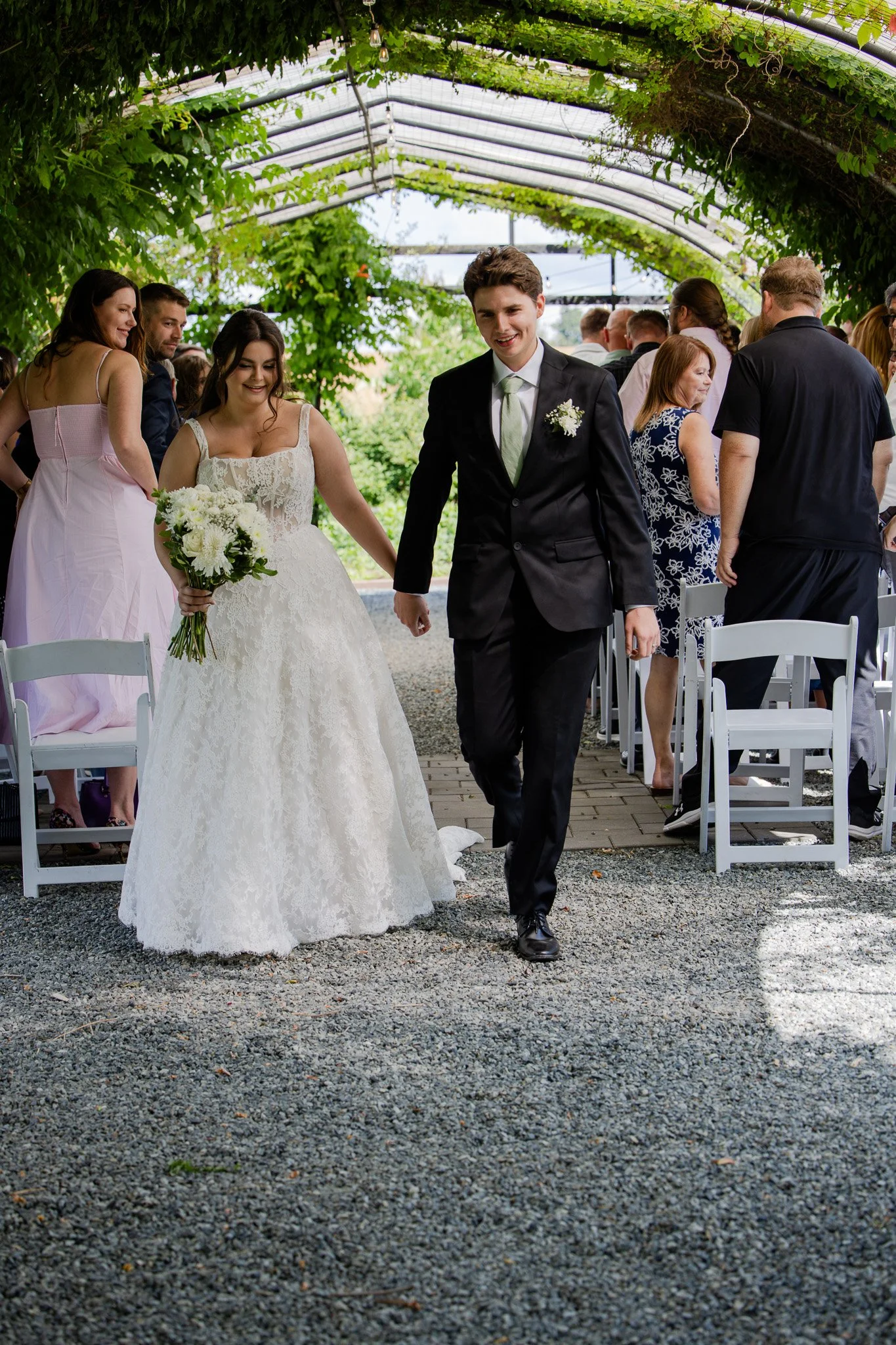 A newlywed couple walking hand in hand at their wedding reception outdoors under a green canopy, with guests mingling in the background.