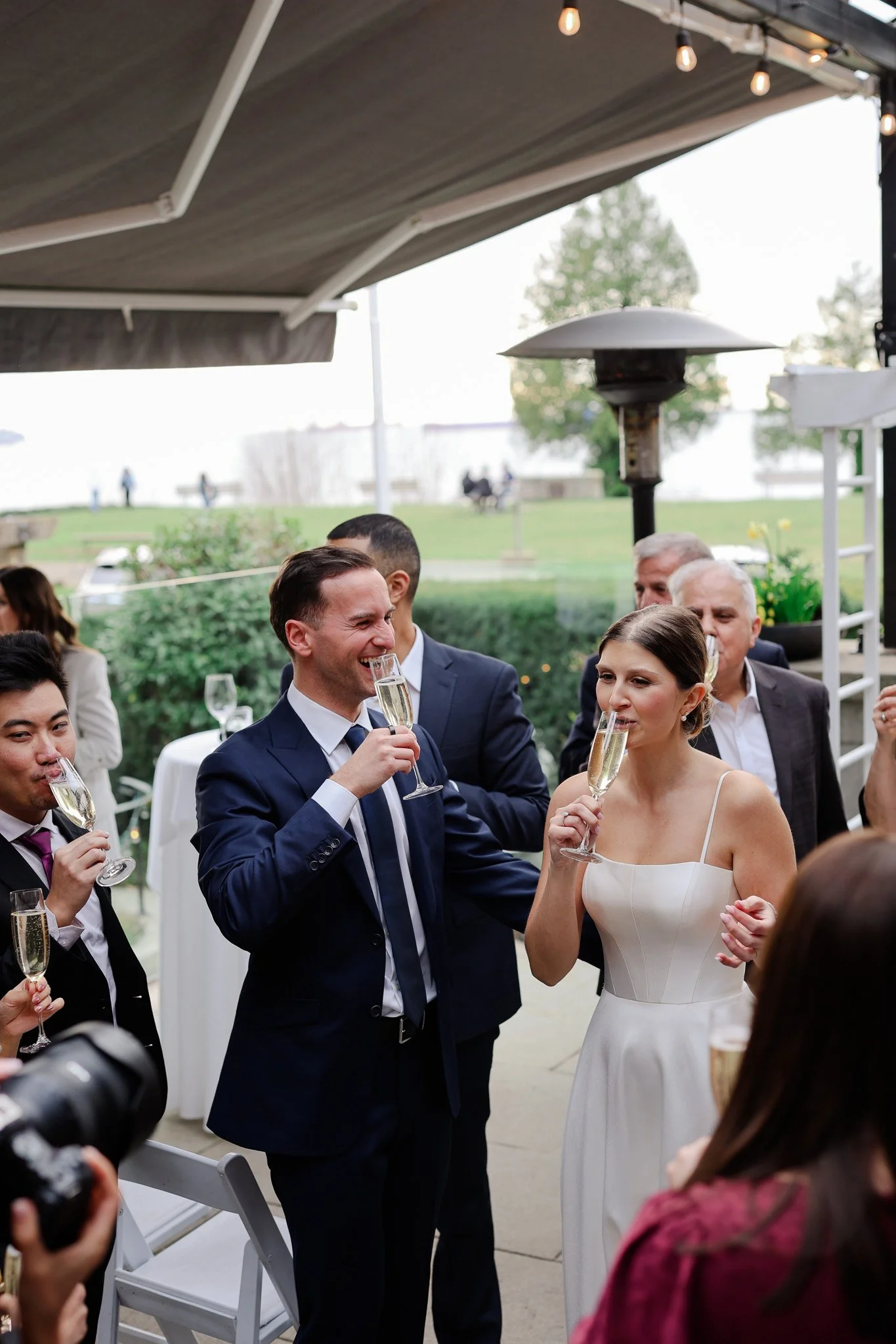 People celebrating at a wedding reception, raising glasses of champagne in toast outdoors under a canopy, with trees and a grassy area in the background.