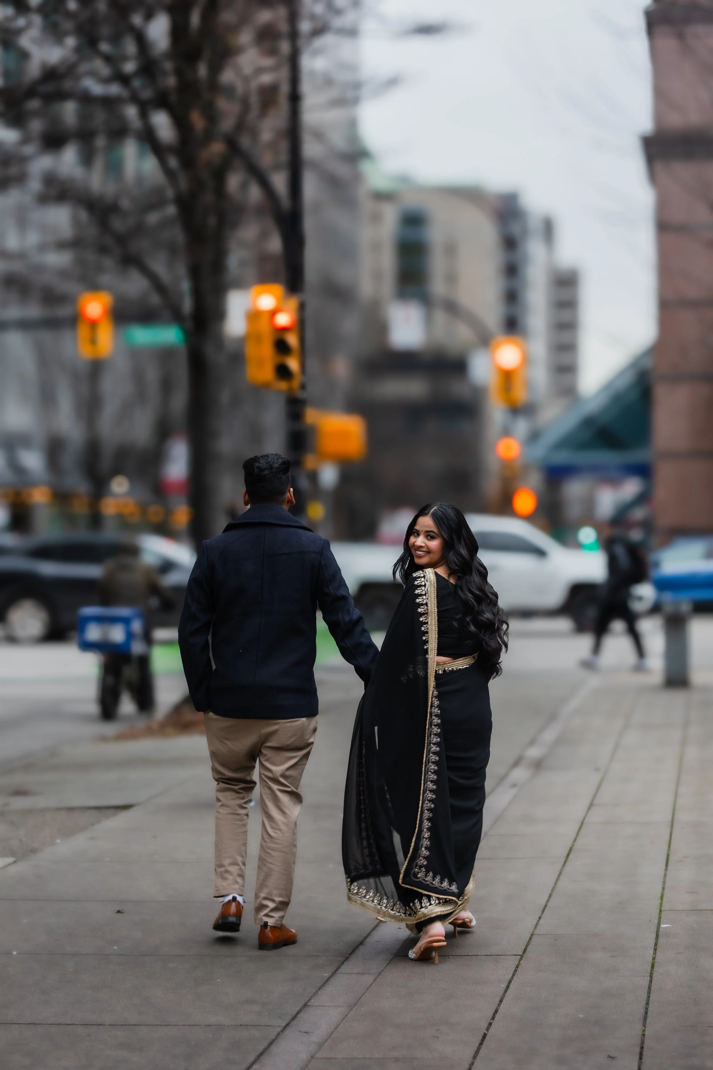 A couple walking on a city sidewalk, woman smiling at the camera, woman wearing a traditional black and gold saree, man in a dark jacket and beige pants, blurred city scene with cars and traffic lights in the background.