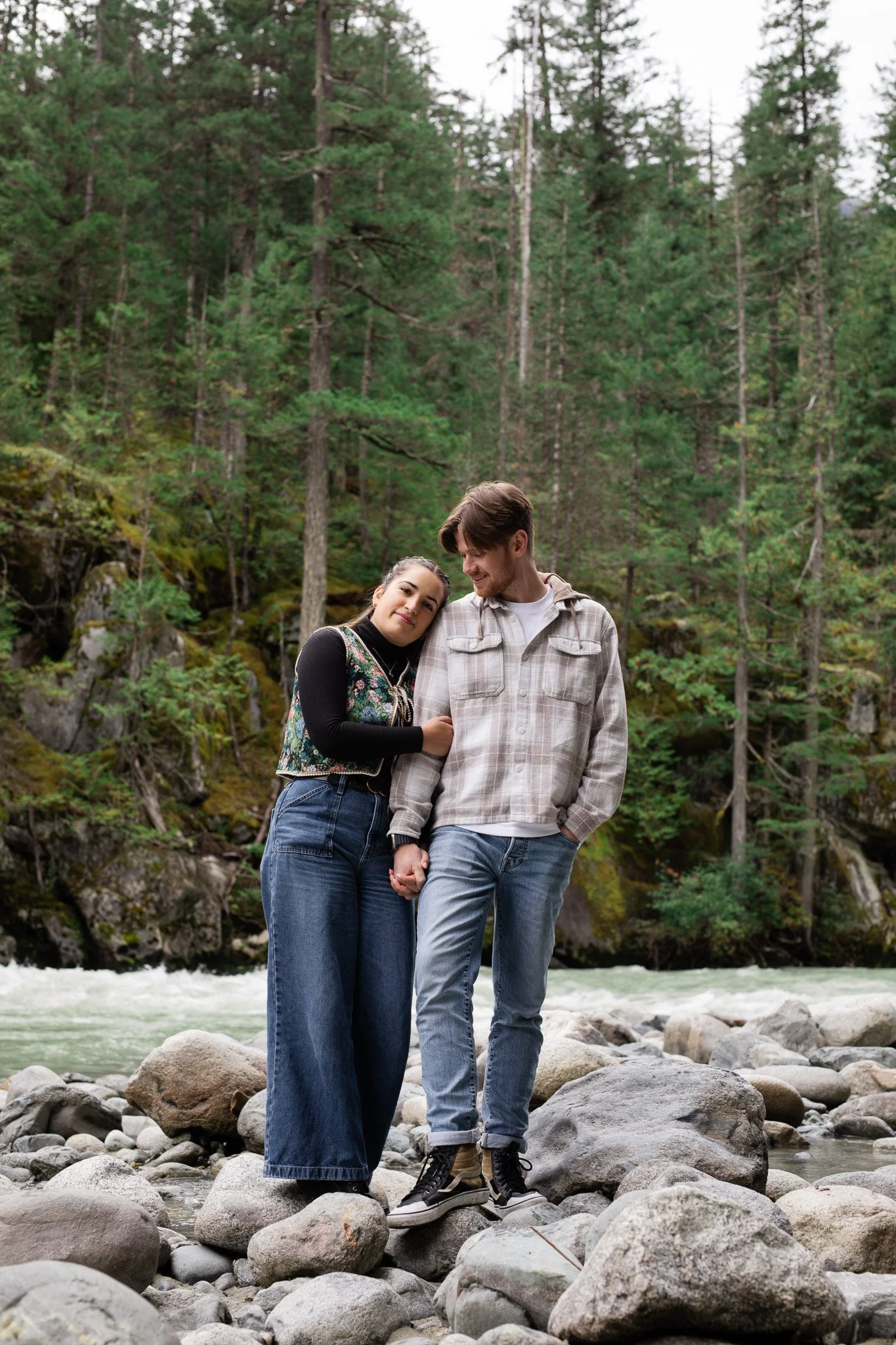 A young couple holding hands and leaning on each other while standing on rocks by a river in a forest.