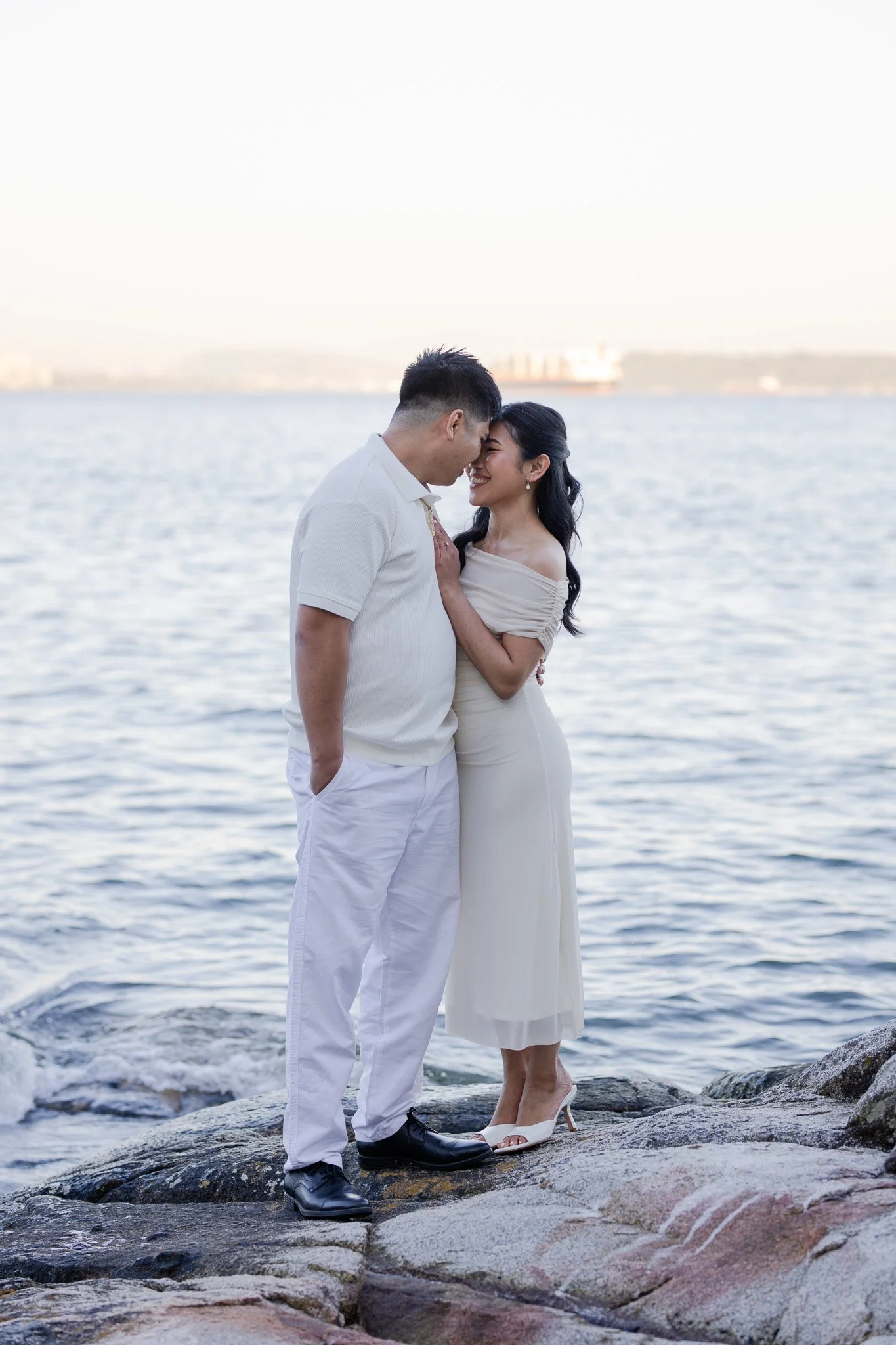 A couple standing close together on rocks near the water, forehead to forehead, in a romantic moment at the seaside during daylight.