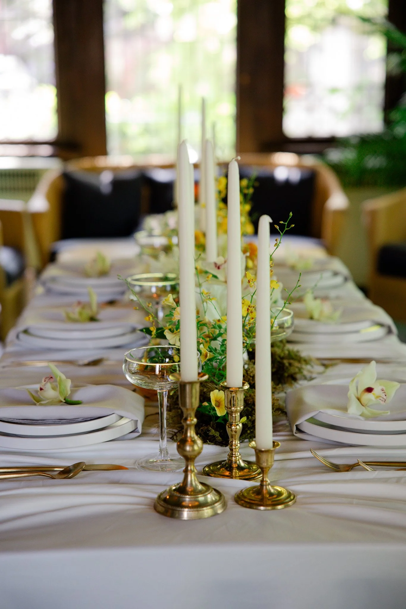 Elegant dining table set with white plates, glasses, and floral decorations, featuring tall white candles in gold candleholders. The table is decorated with flowers and has a soft, natural light coming through large windows in the background.