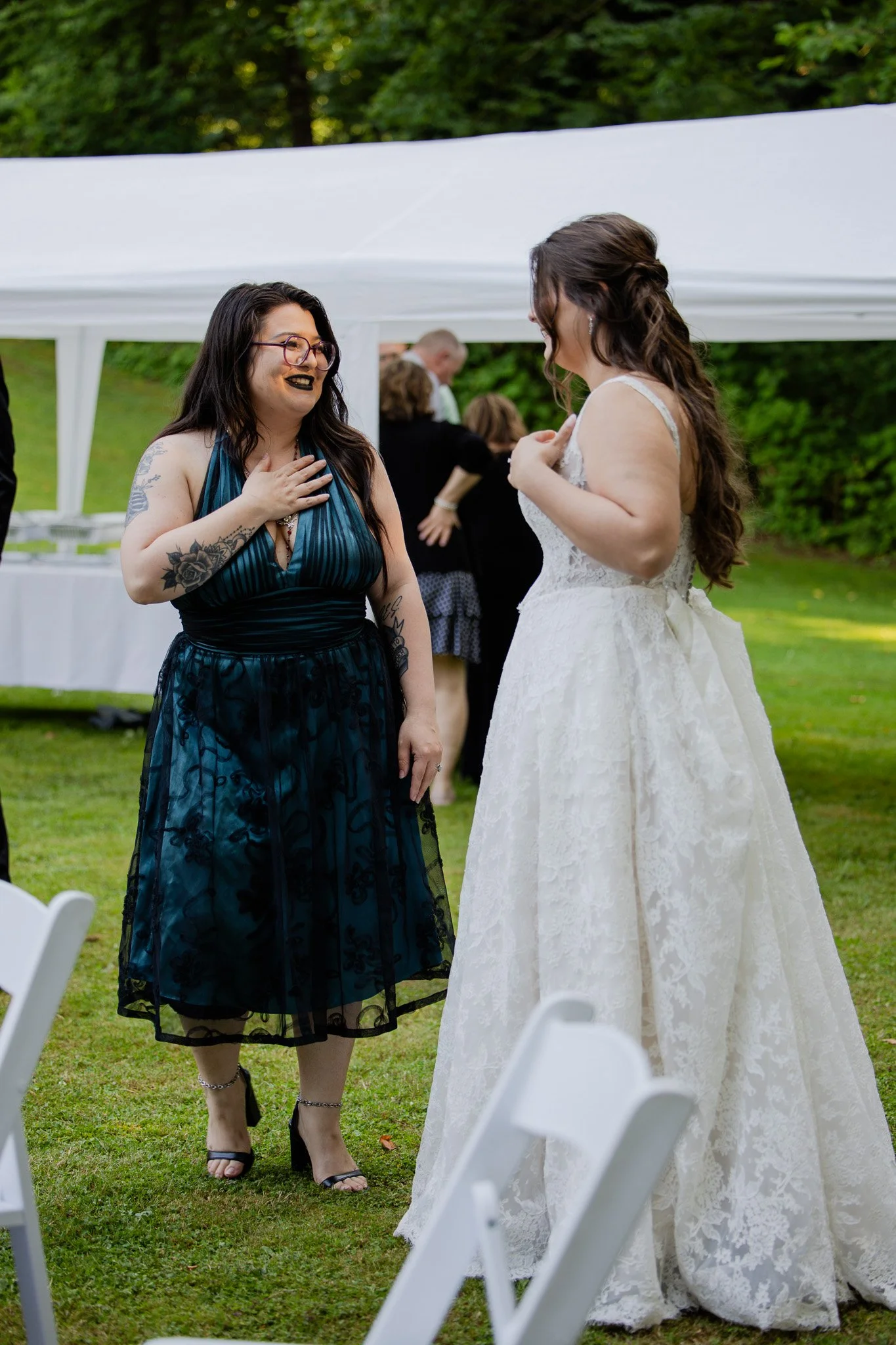 Two women at a wedding, one in a white wedding dress and the other in a dark blue dress, smiling and engaging in conversation outdoors under a white canopy, with green trees in the background.
