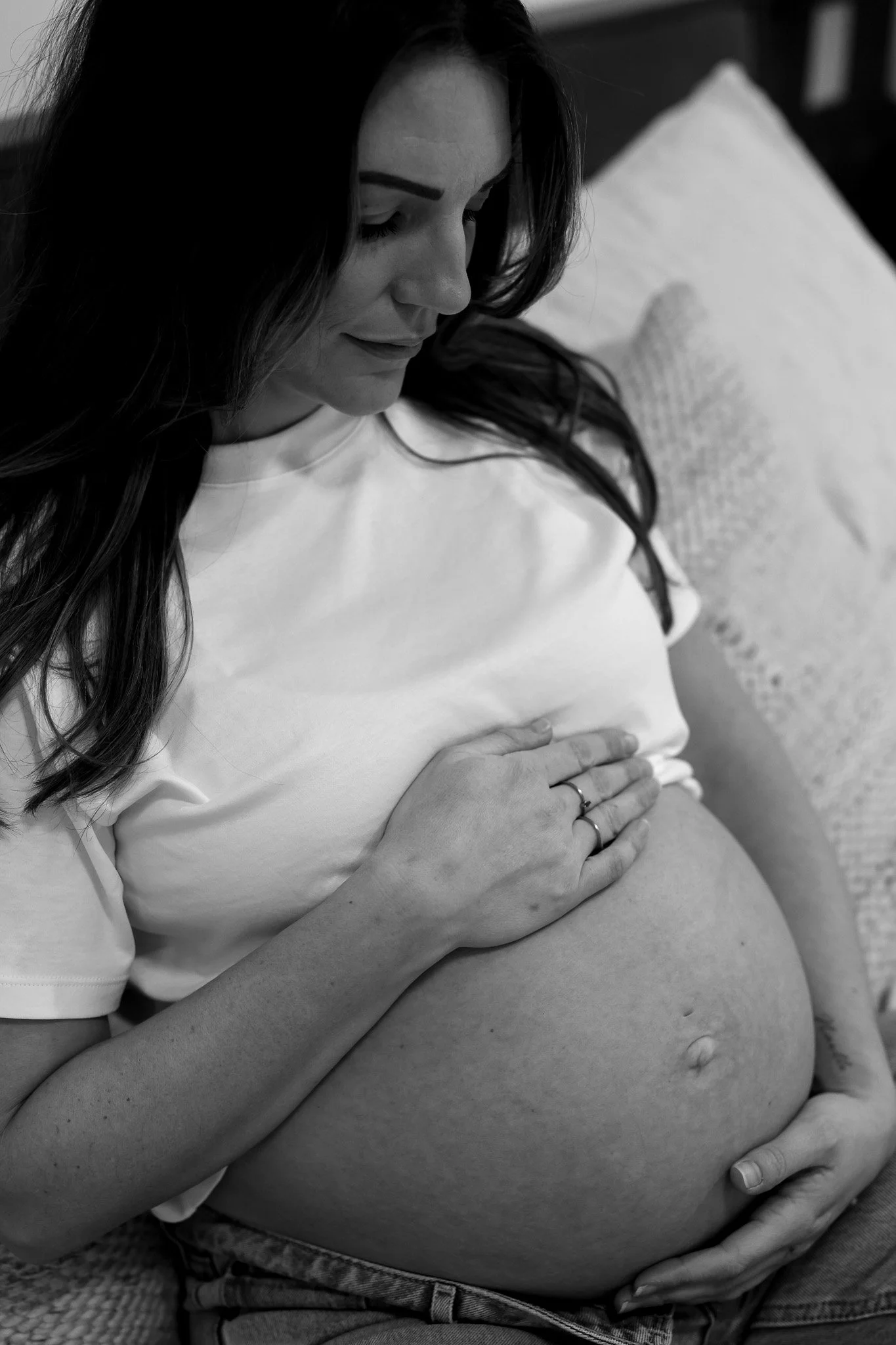 A pregnant woman sitting on a bed, gently touching her belly and smiling with her eyes closed.
