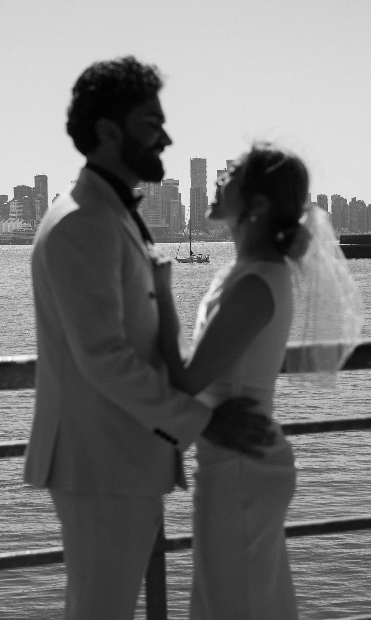 Silhouette of a couple holding hands and looking at each other by the water, with city skyline and sailboat in the background.