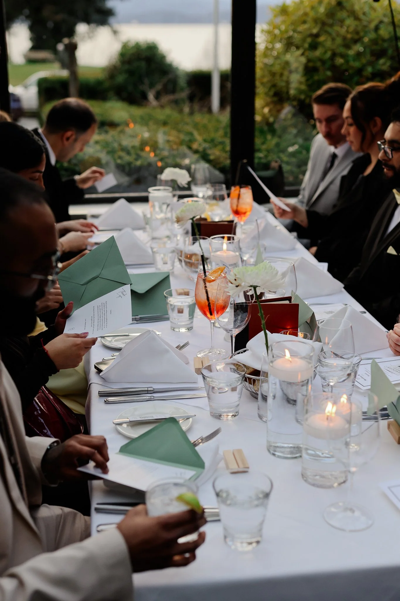 People seated at a formal dining table during a gathering or celebration, with candles, flowers, drinks, and menus on the table, in a bright room with large windows showcasing an outdoor view.