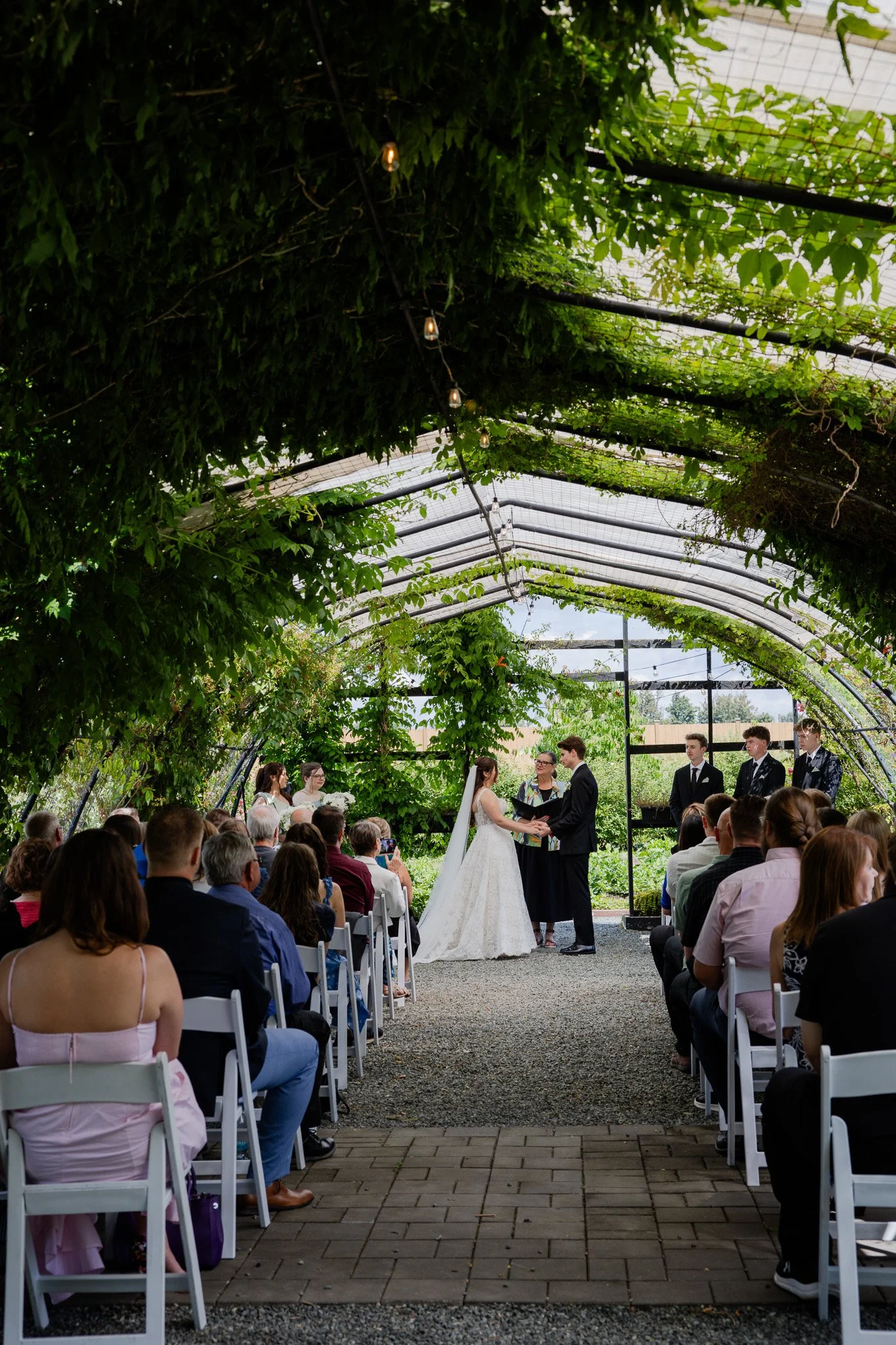 A wedding ceremony taking place in a greenhouse with lush green foliage overhead. The bride and groom hold hands and exchange vows, surrounded by seated guests. The image captures a moment of intimacy and celebration.