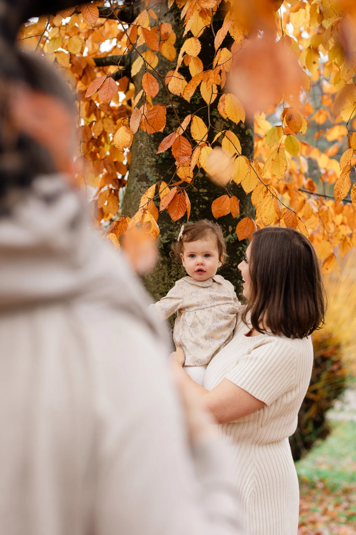 A woman holding a young girl outdoors during autumn, with orange and yellow leaves on a large tree in the background.