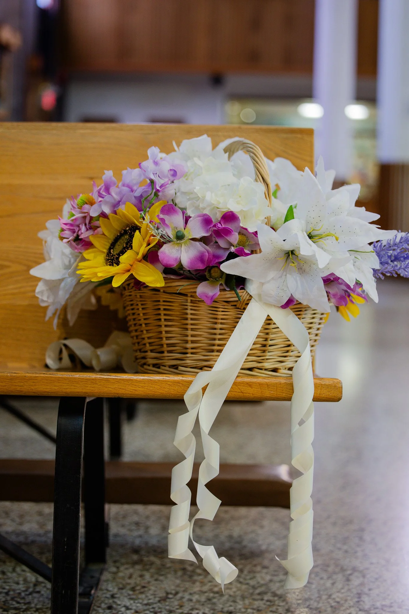 A basket of artificial flowers, including white lilies, purple orchids, yellow daisies, and other colorful blossoms, with white ribbon streamers, placed on a wooden table.