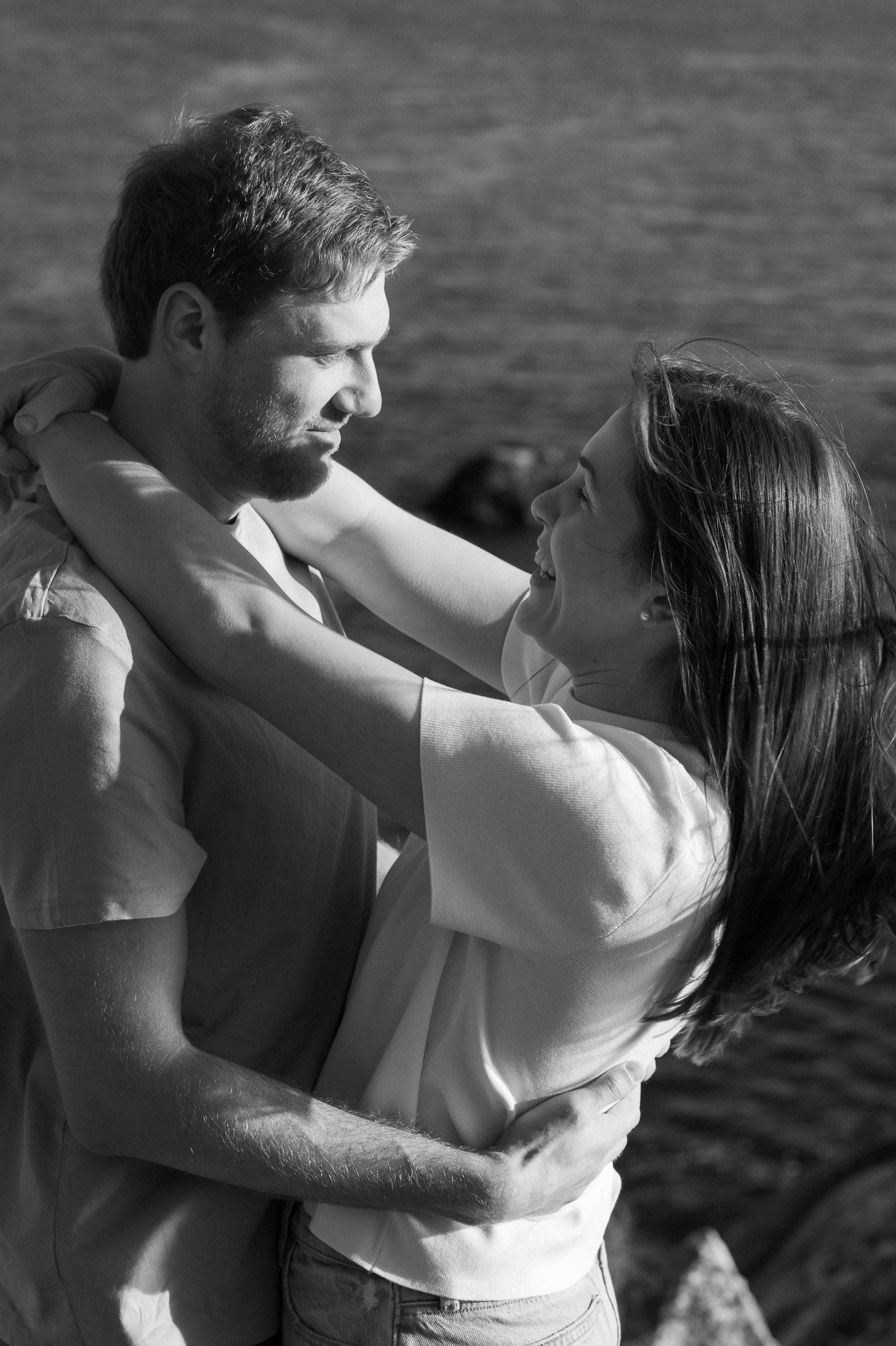A smiling couple embracing near a body of water, with the man holding the woman close and both looking at each other lovingly.