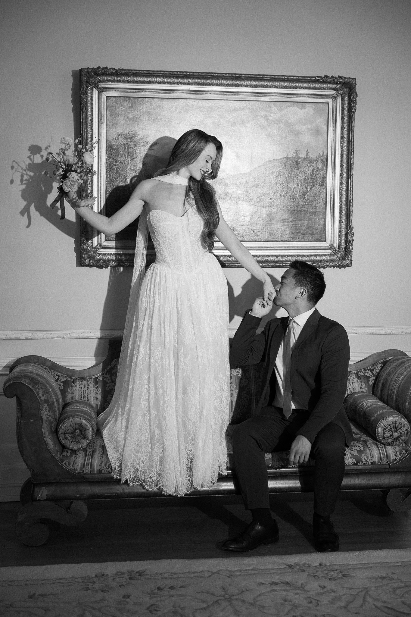 A black and white photo of a woman in a wedding dress standing on a vintage sofa, holding a bouquet and smiling. A man in a suit is sitting on the sofa, kissing her hand. The background features a landscape painting framed on the wall.