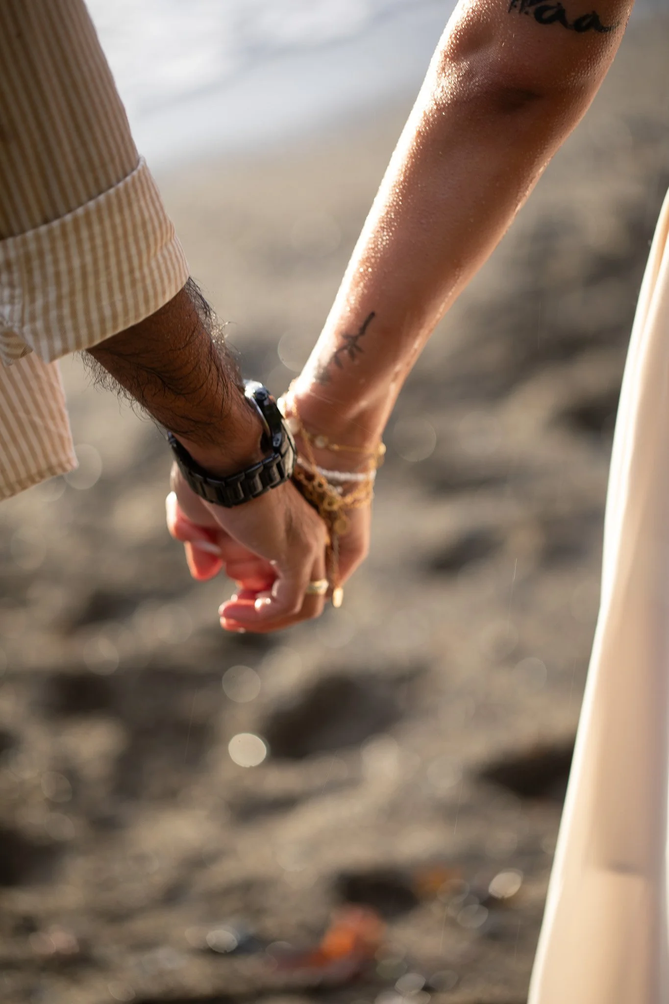 Close-up of two people holding hands, one with a watch and bracelets, the other with a tattoo, on a sandy beach.