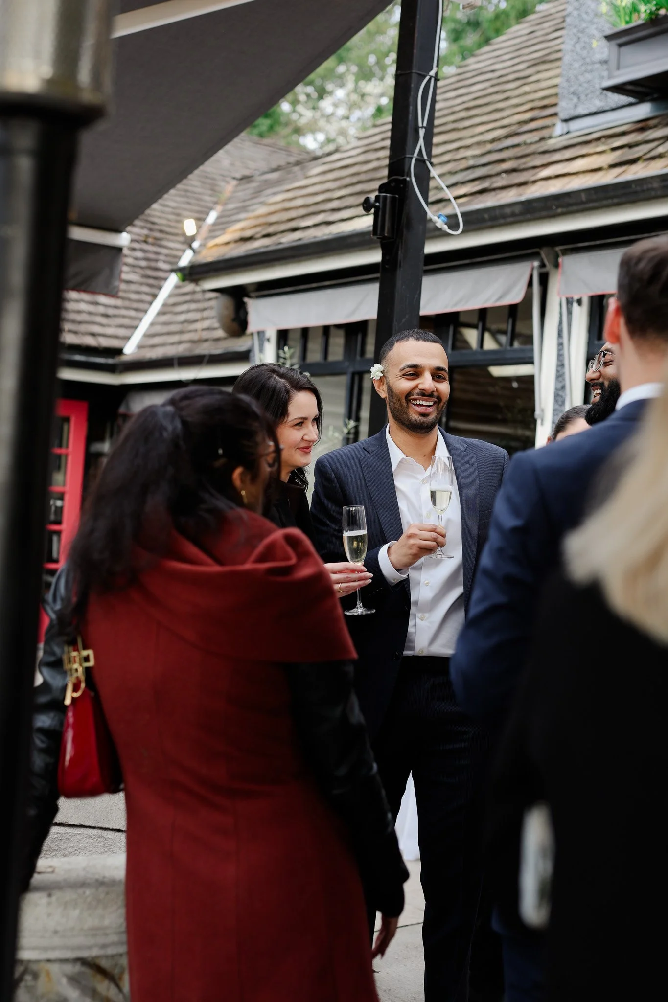 Group of people gathered outdoors, socializing and holding champagne glasses, with a man smiling wearing a suit and white dress shirt, a woman with dark hair and a flower in her hair, and others partially visible.