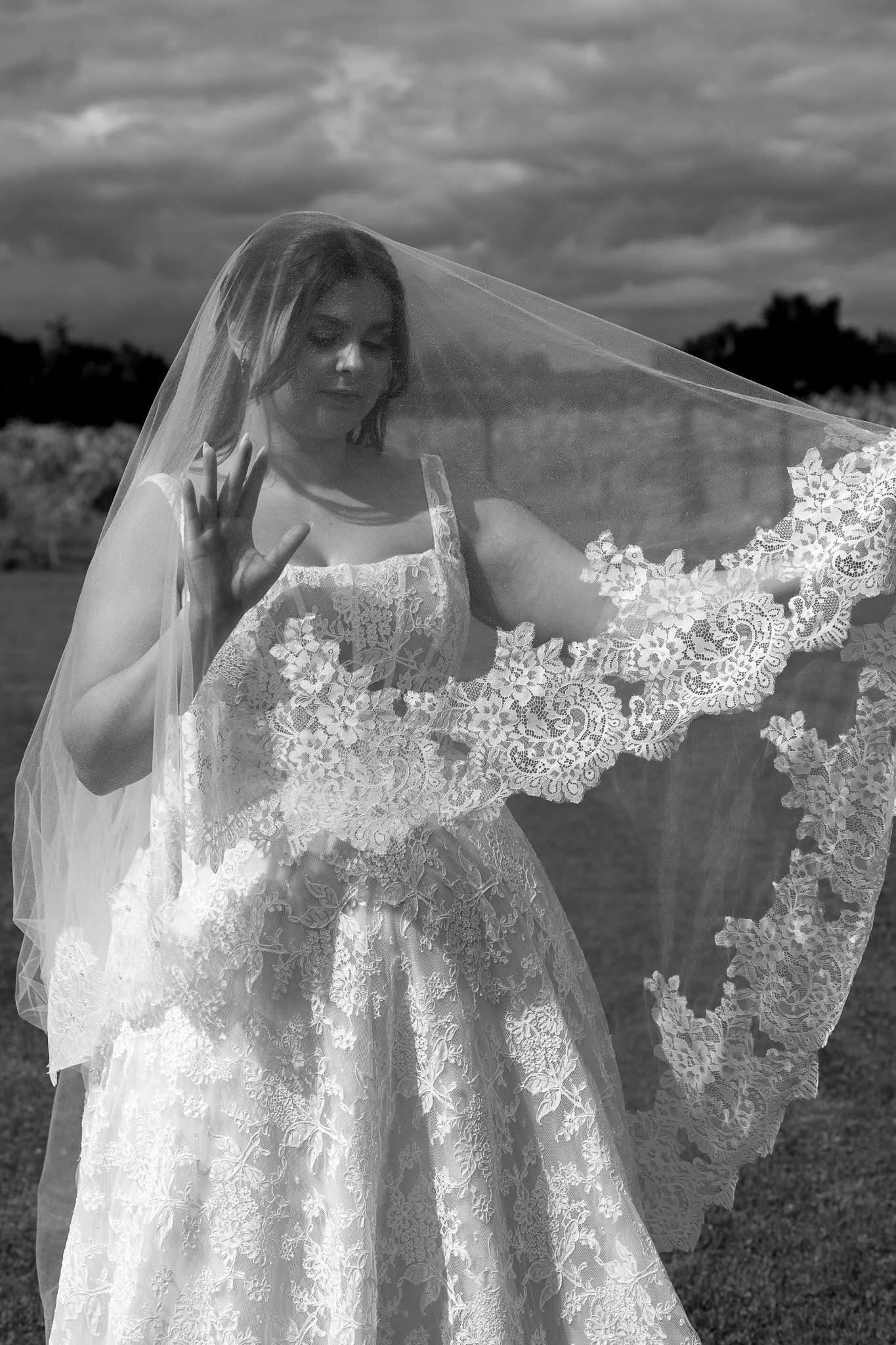A woman in a wedding dress and veil standing in an open field with trees in the background, outdoors during the daytime, in black and white.