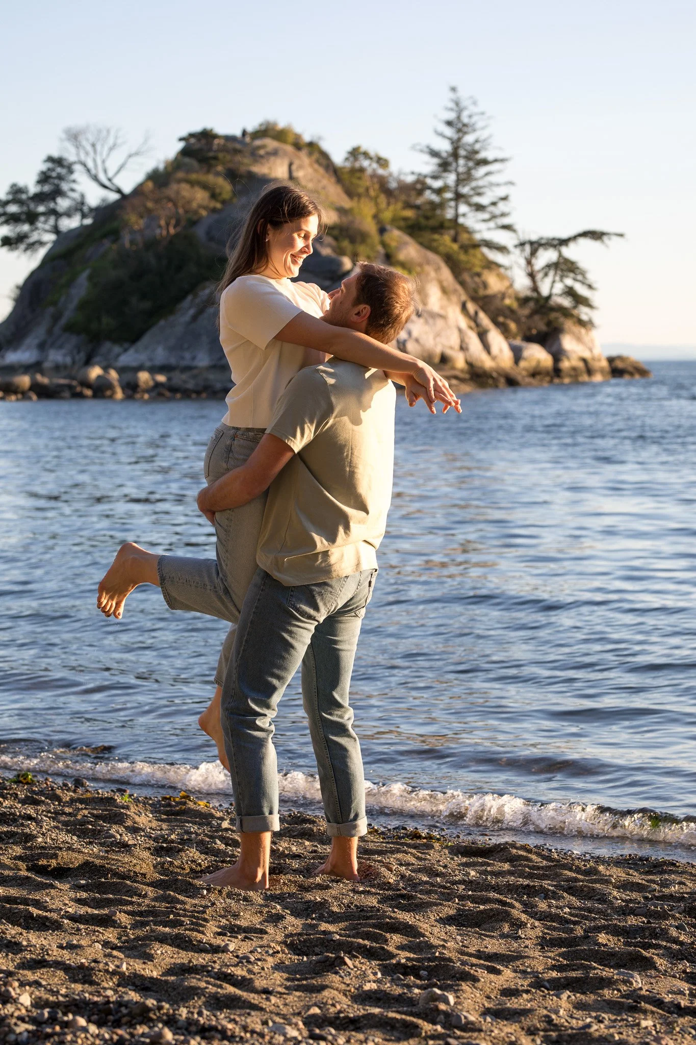 A couple at the beach, with the woman sitting on the man's lap, both smiling, near a rocky outcrop with trees, during sunset.