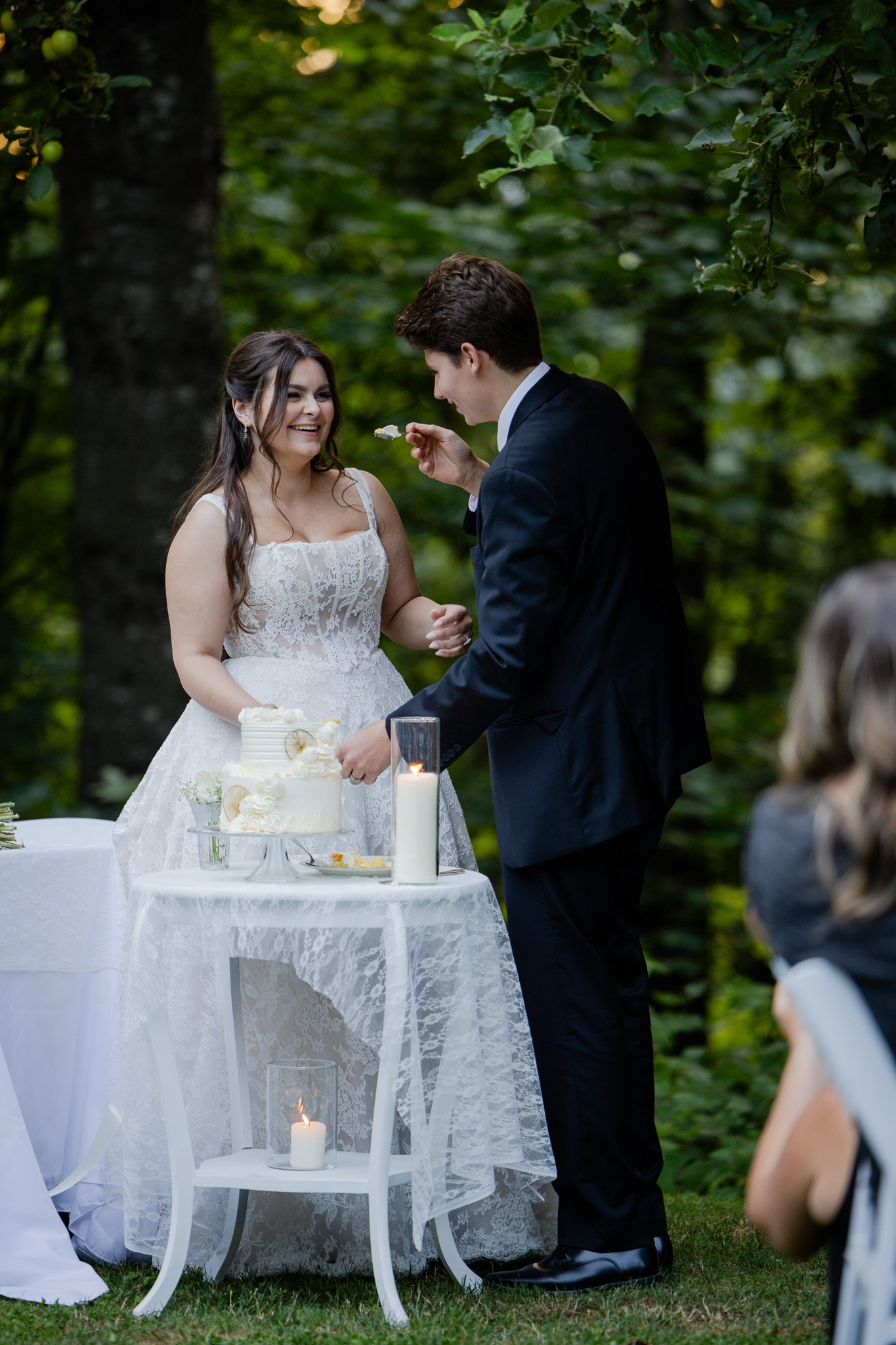 A couple at their wedding, standing outside in a wooded area. The bride is in a white lace wedding dress, smiling, as the groom feeds her cake. There is a wedding cake on a white lace-covered table, along with candles and glassware.