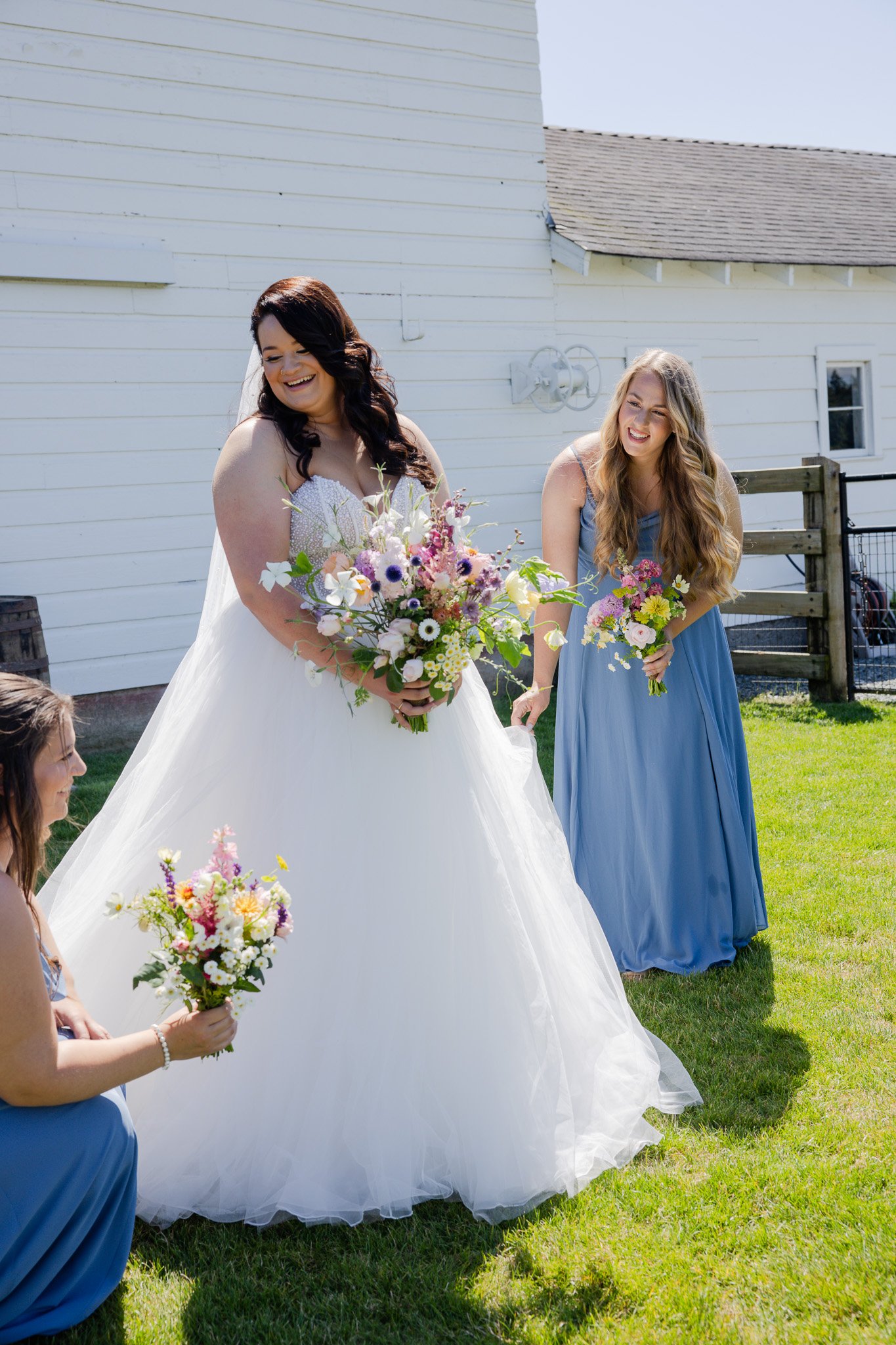 A bride in a white wedding gown holding a bouquet of flowers, smiling, with a bridesmaid in a blue dress holding a bouquet, outdoors on grass near a white building.