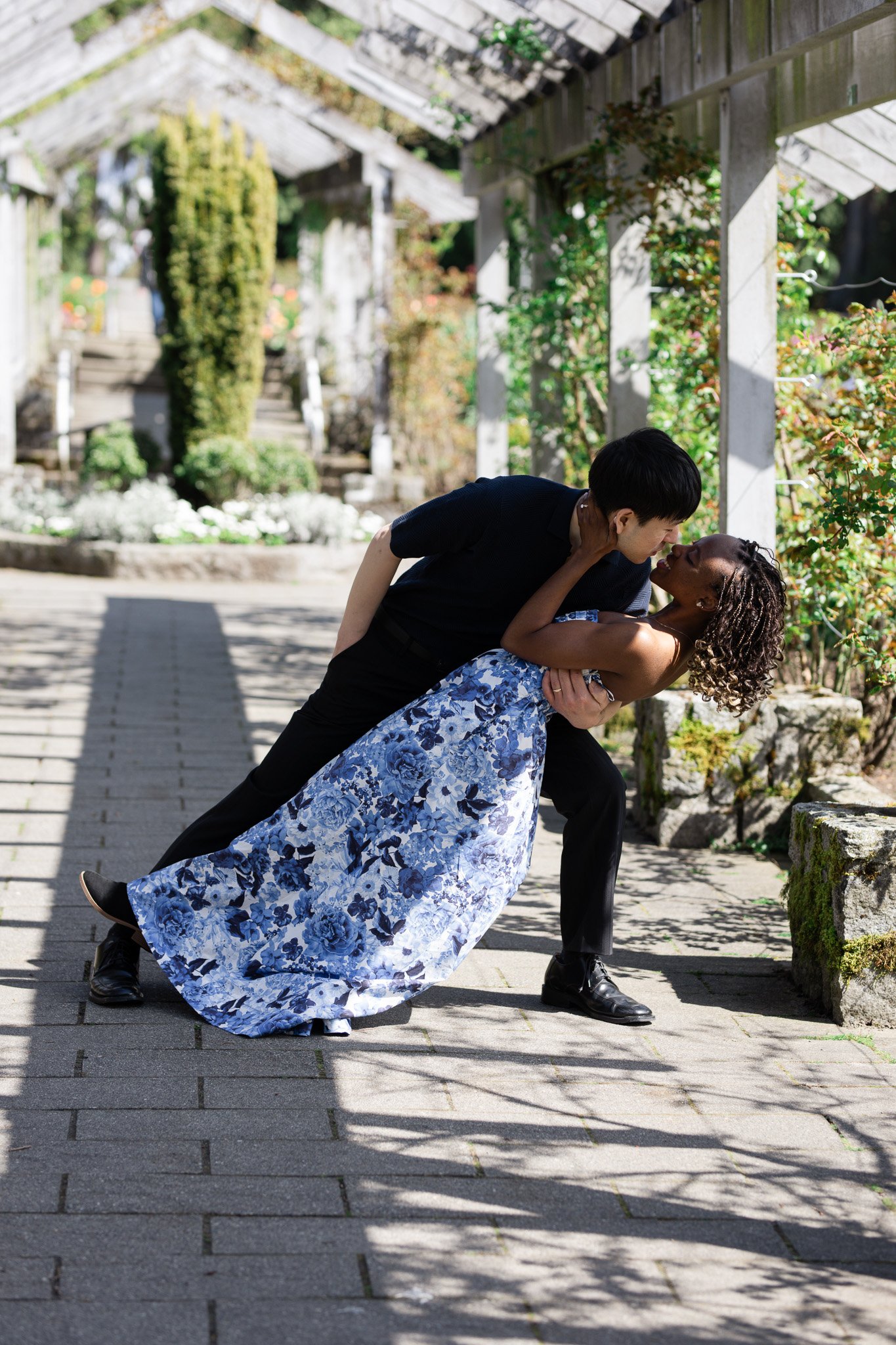 A man in black clothing dips a woman in a blue floral dress in an outdoor garden setting, both smiling and about to kiss.