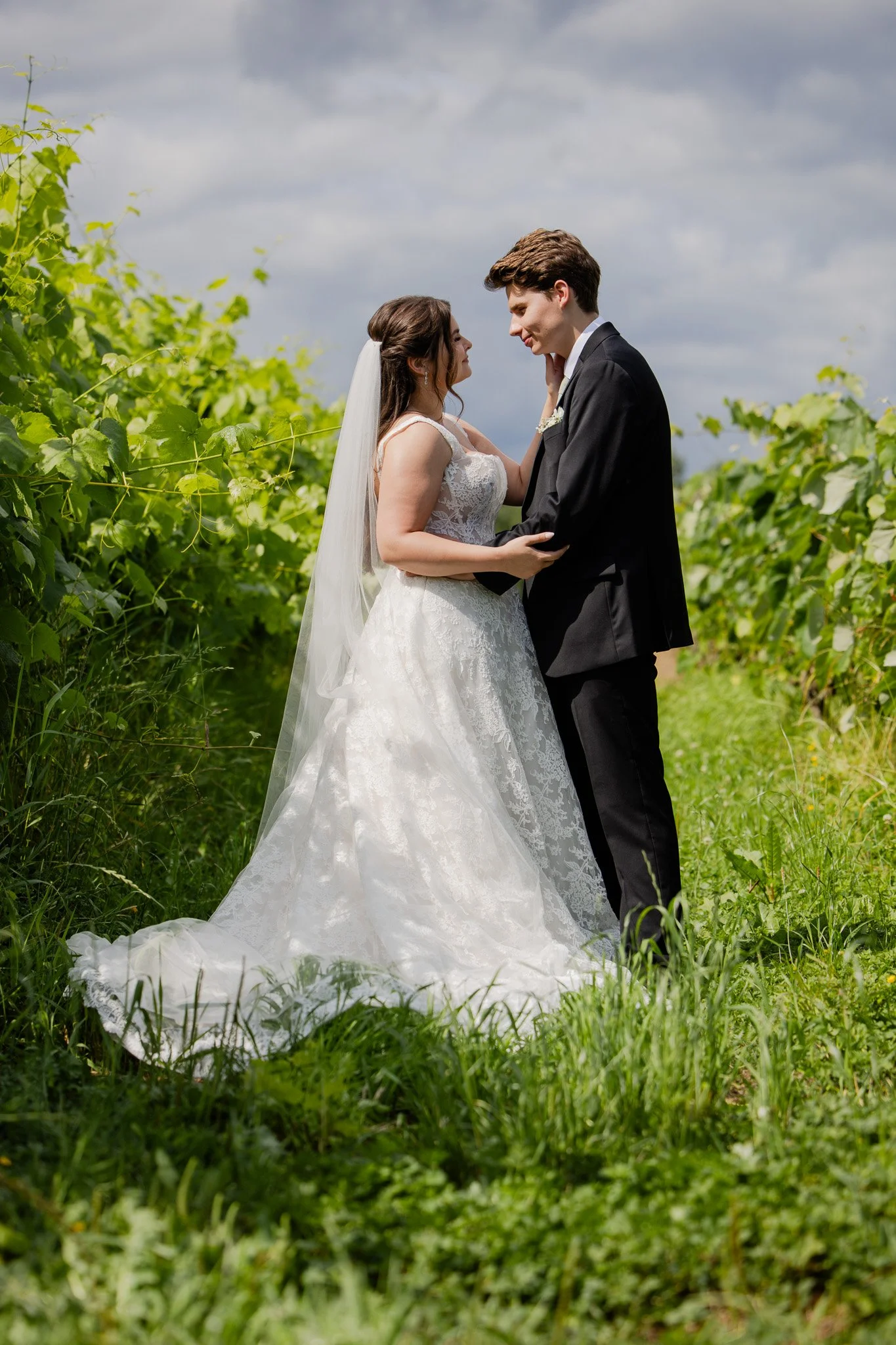 A bride and groom embracing outdoors among green foliage on a cloudy day.