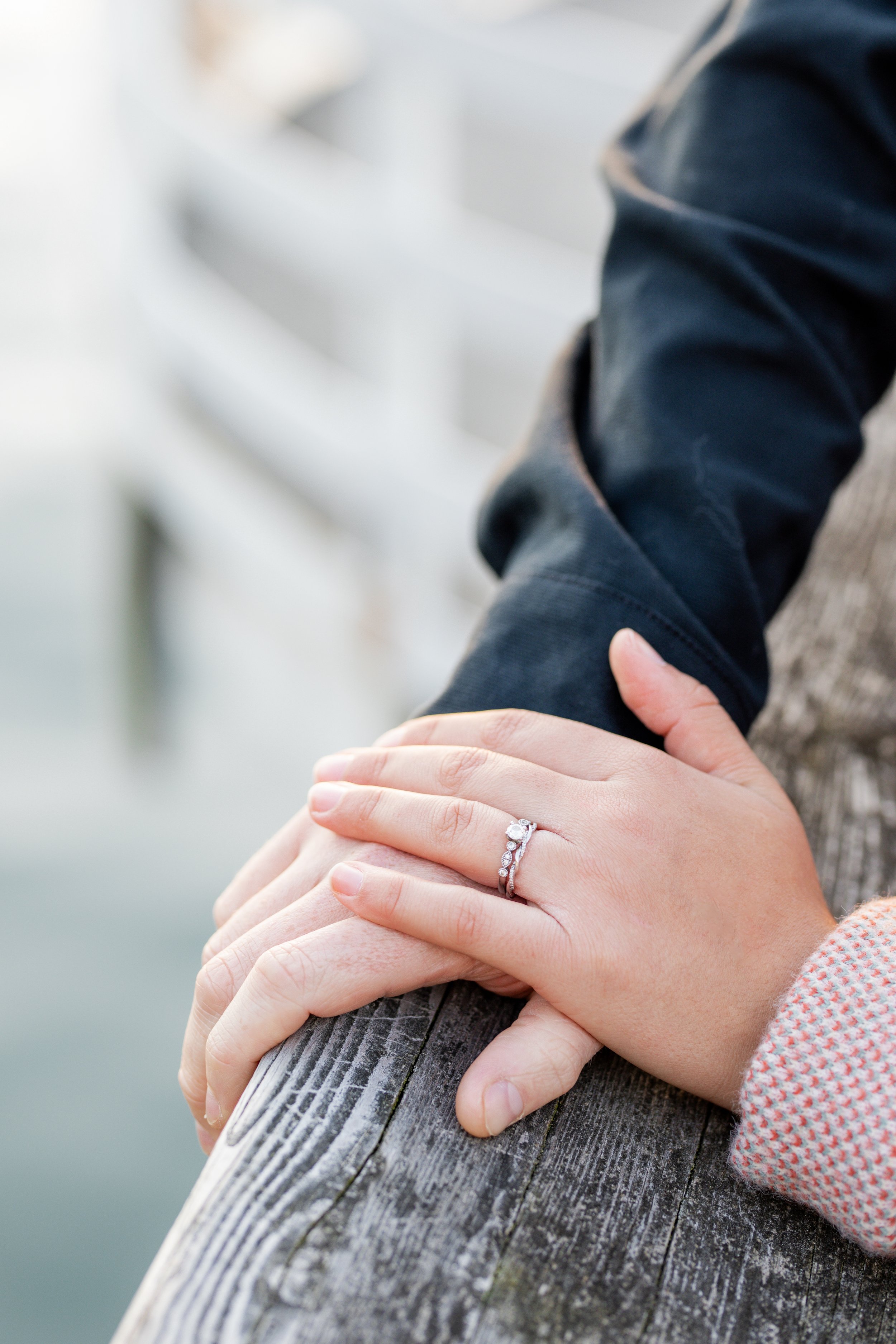 Close-up of a woman's hand with an engagement ring resting on a man's hand, holding a wooden railing outdoors near water.
