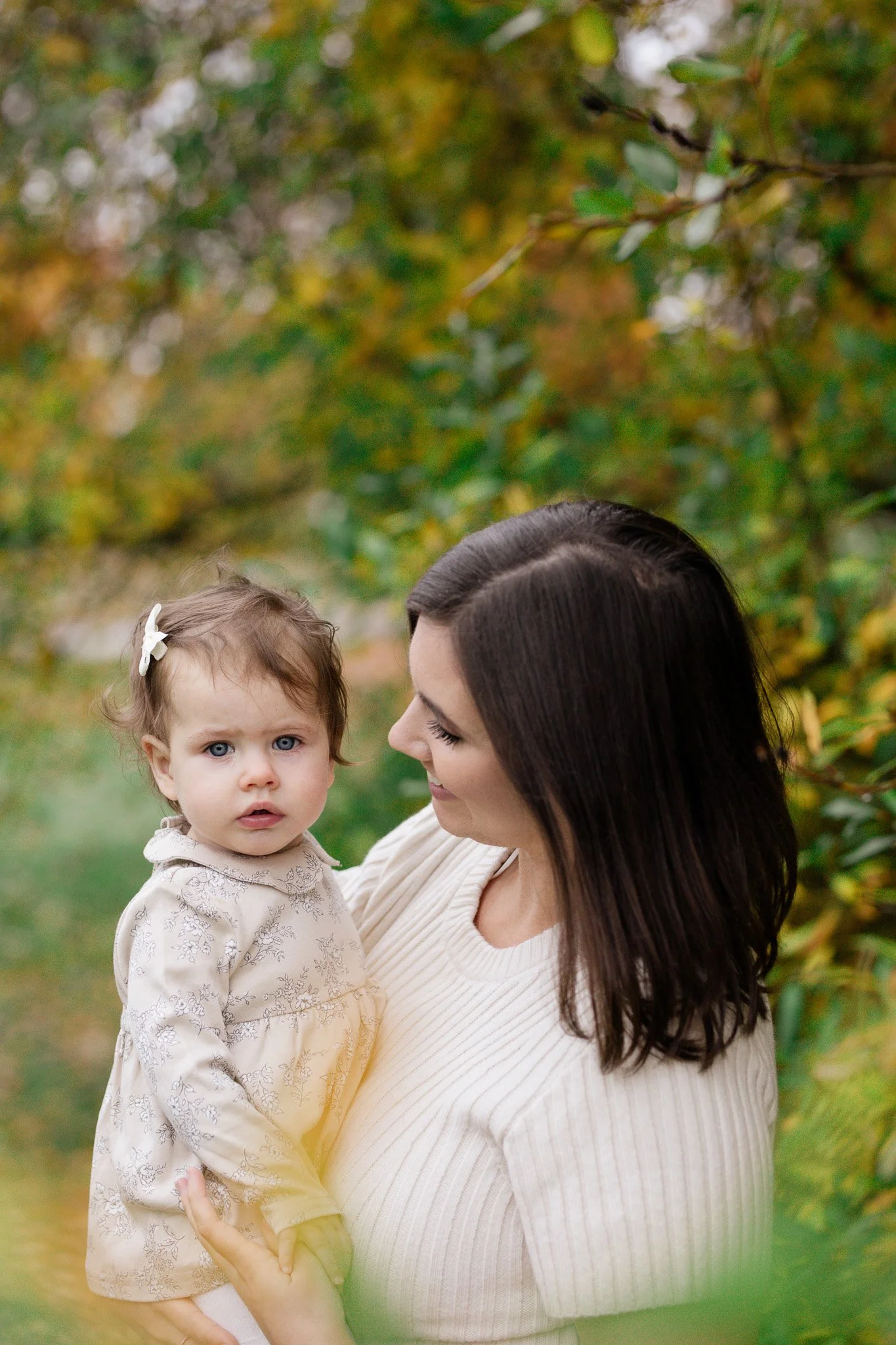 A woman holding a young girl outdoors among autumn trees, with fall foliage in the background.