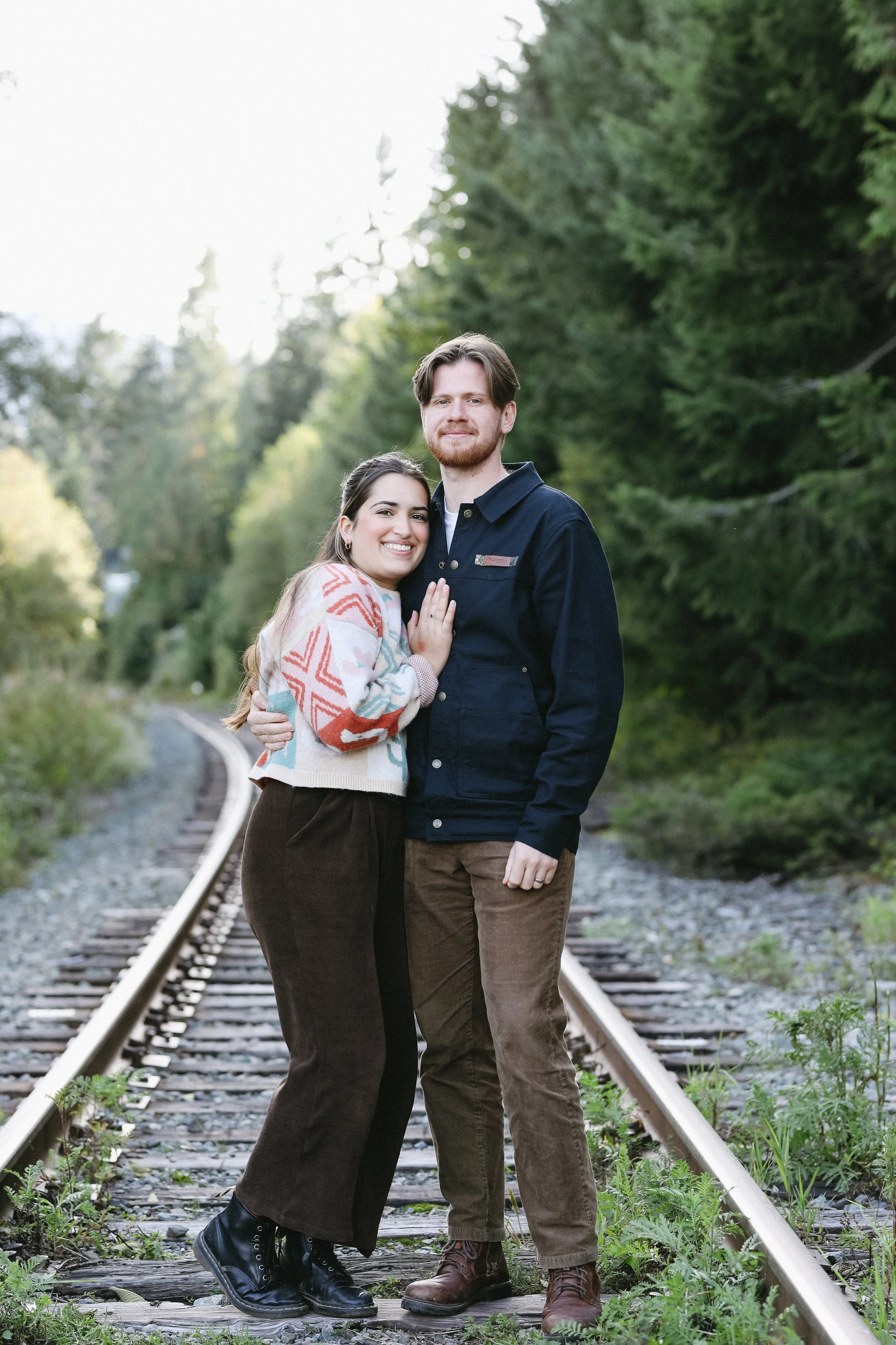 A happy couple standing on a railway track surrounded by greenery, holding each other and smiling at the camera.