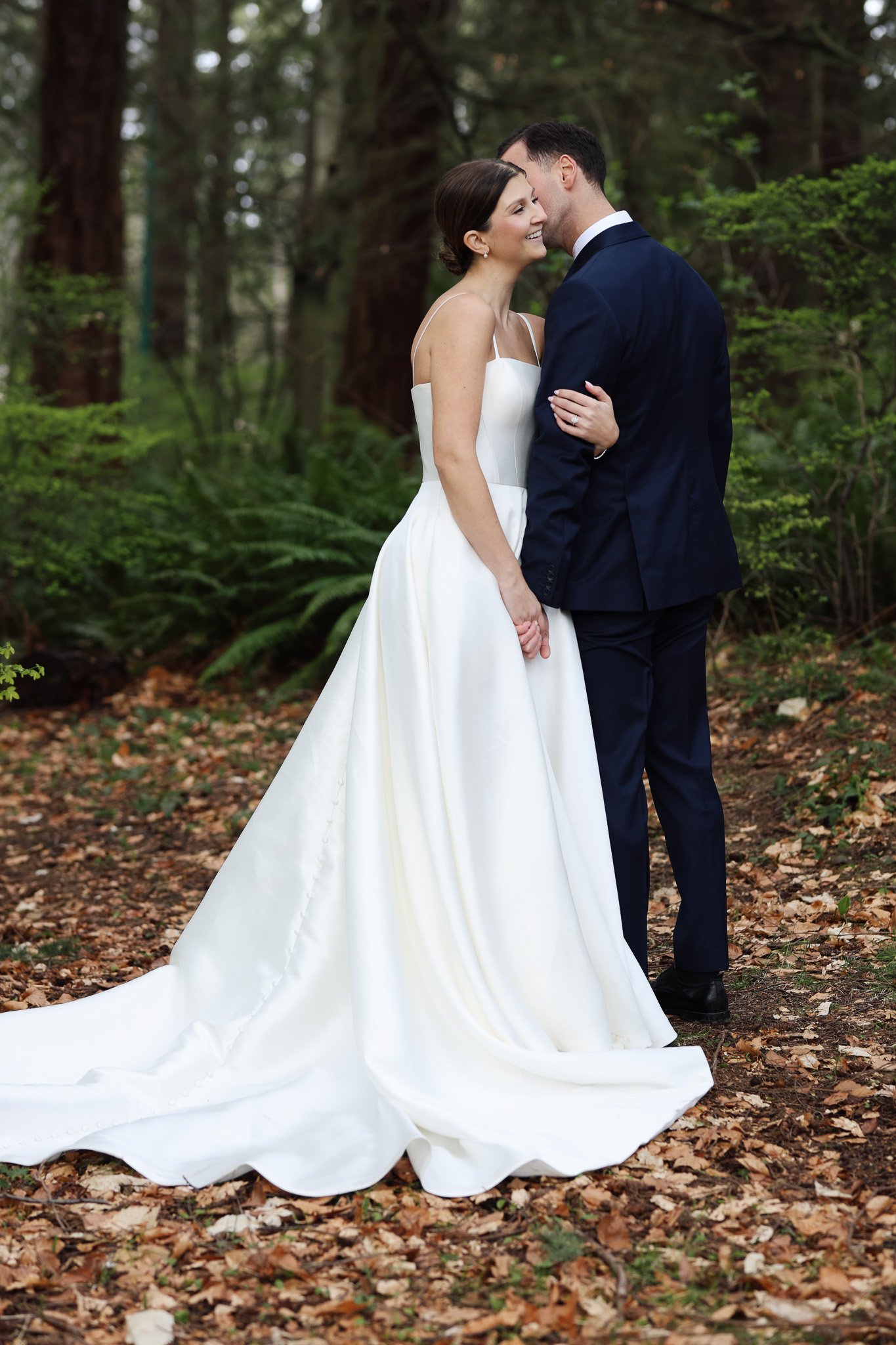 A smiling bride and groom sharing a tender moment in a lush forest setting, with fallen leaves and greenery surrounding them.
