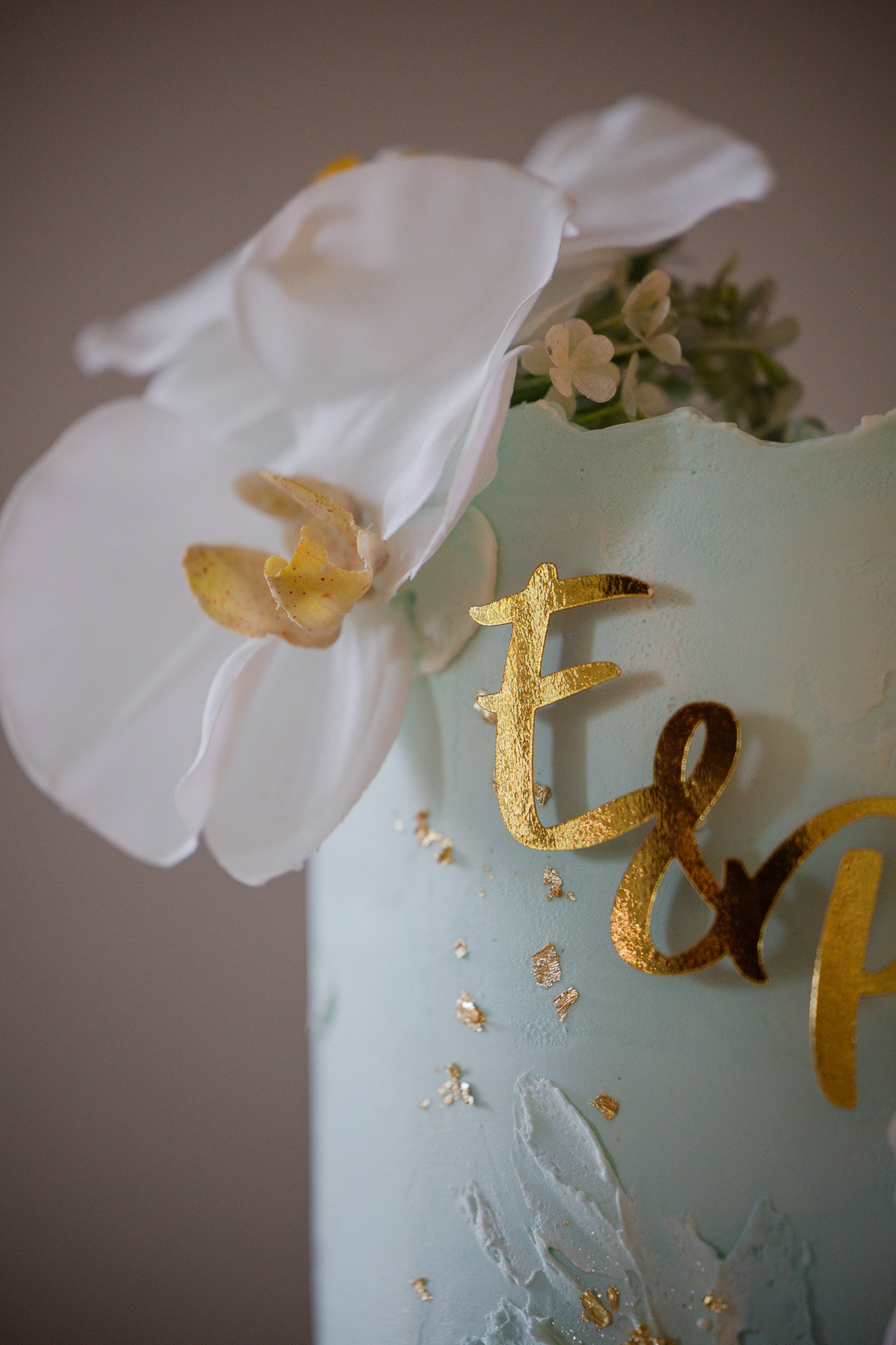 Close-up of a decorated cake with white icing, white flowers, gold lettering, and small gold leaf accents.
